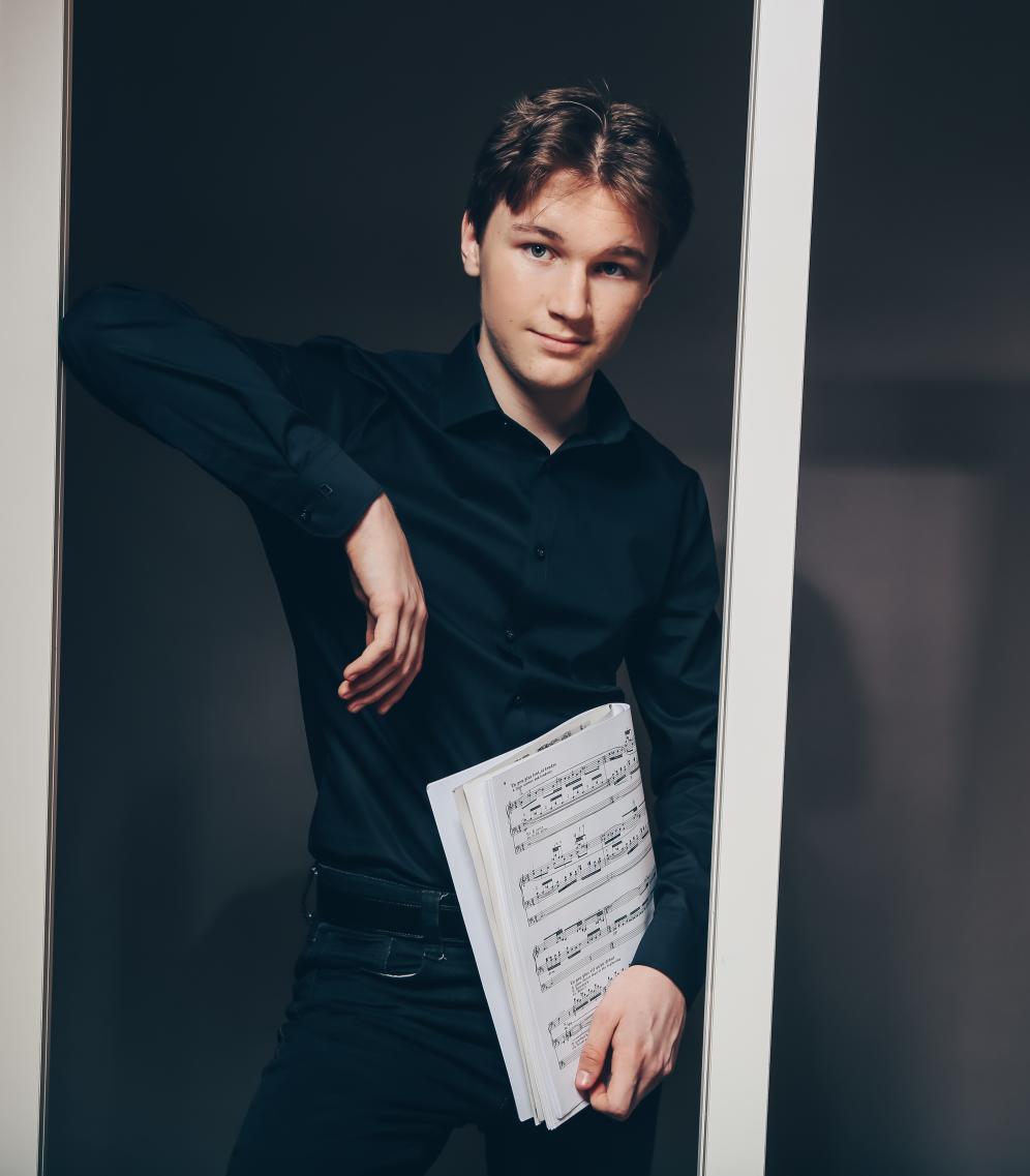 Junger Mann mit Noten in der Hand. Er lehnt sich an einen weißen Rahmen.