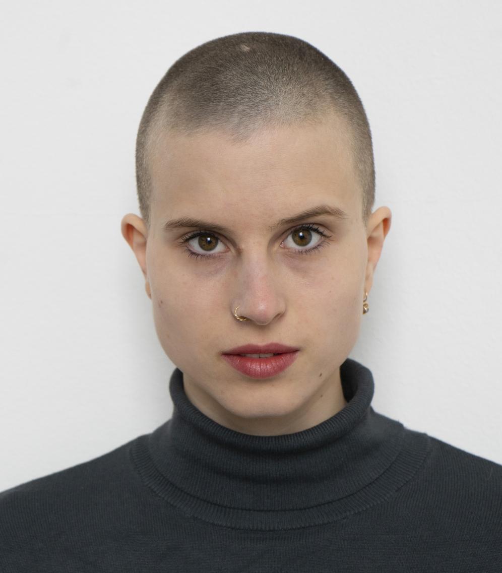 Das Bild zeigt einen Headshot von Lisa Freiberger. Sie isst dezent geschminkt, trägt die Haare in einem Buzzcut und hat einen dunkelgrünen Rollkragenpullover an. 