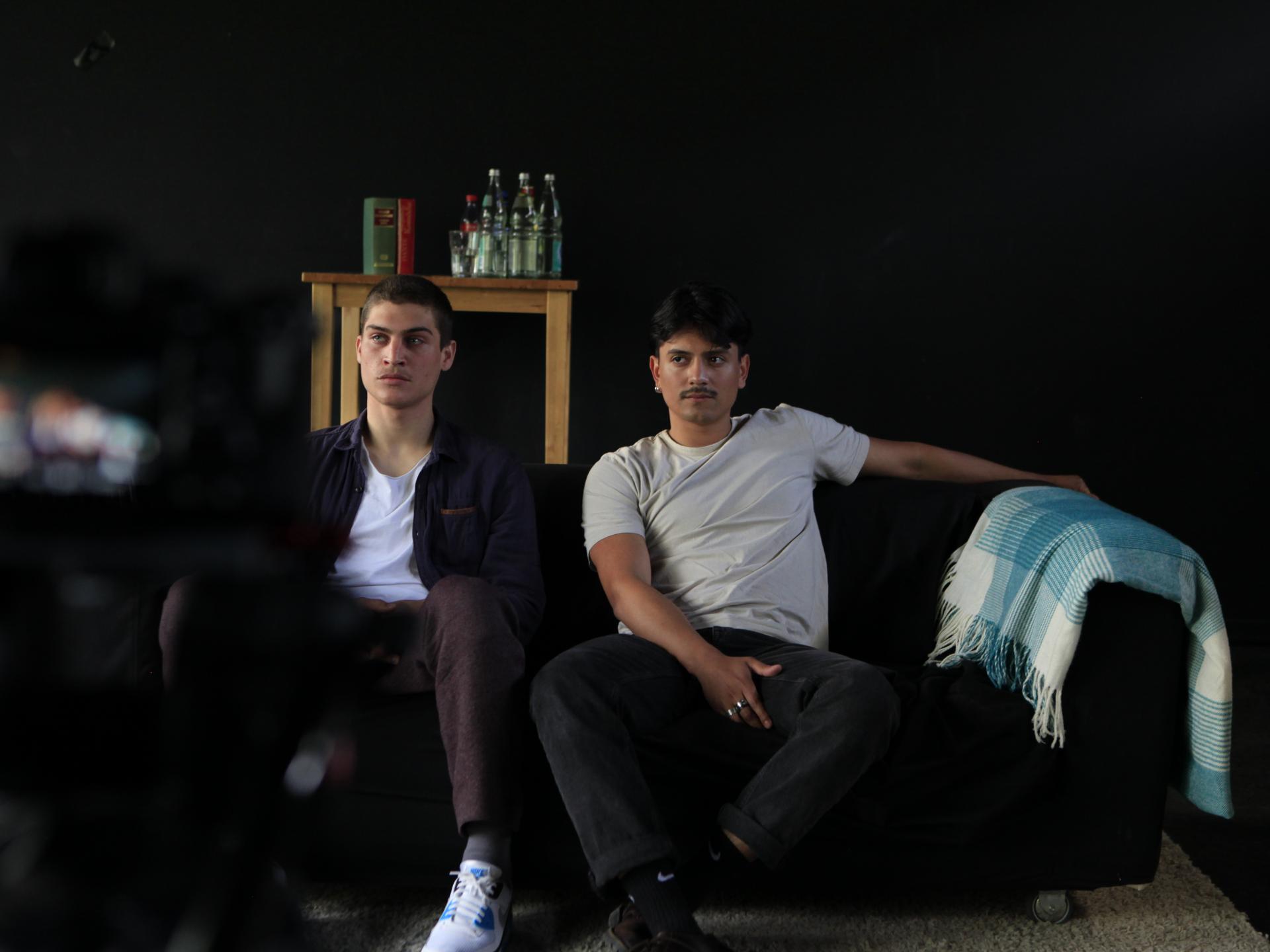 Zwei Schauspielstudierende auf schwarzem Sofa vor einer Filmkamera.