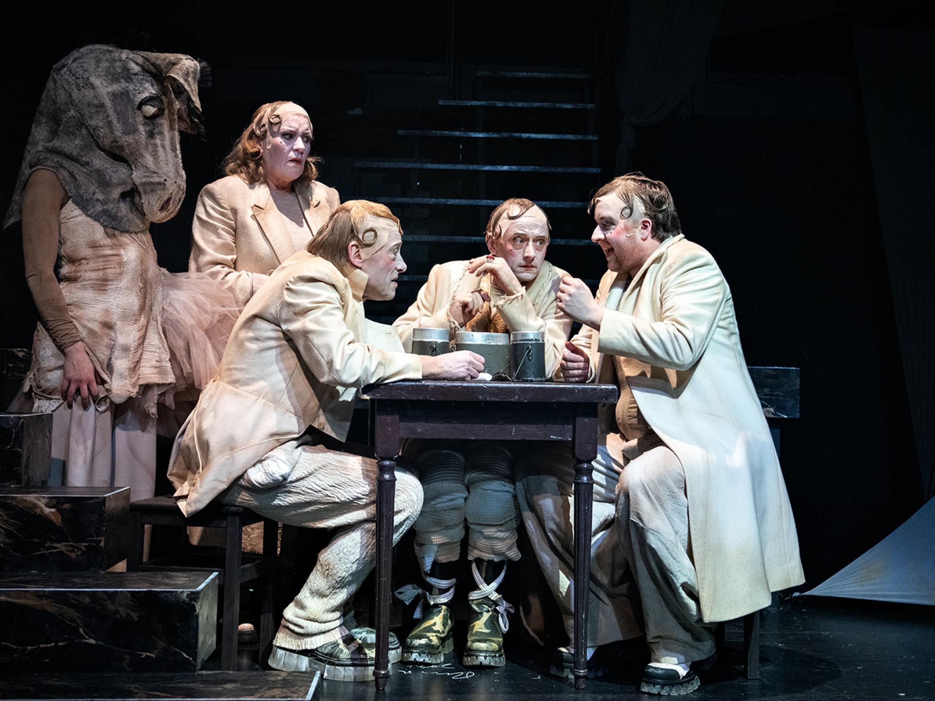 Um einen Tisch herum sitzen bzw. stehen Figuren des Stückes: Tyll (gespielt von Esra Schreier), Agneta, Claus, Kircher und Tesimond in der Inszenierung von "Tyll" am Theater Heidelberg im Dezember 2021.