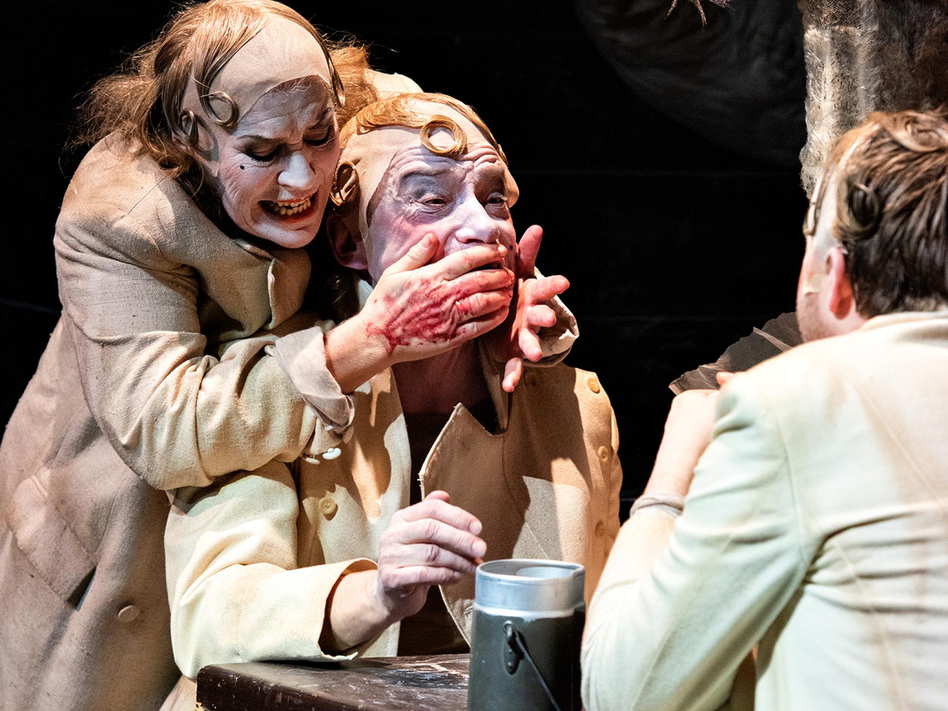 Die Figur Angeta (gespielt von Elisabeth Auer) hält Claus den Mund zu, der an einem Tisch gegenüber von Tesimond sitzt, in der Inszenierung von "Tyll" am Theater Heidelberg im Dezember 2021.