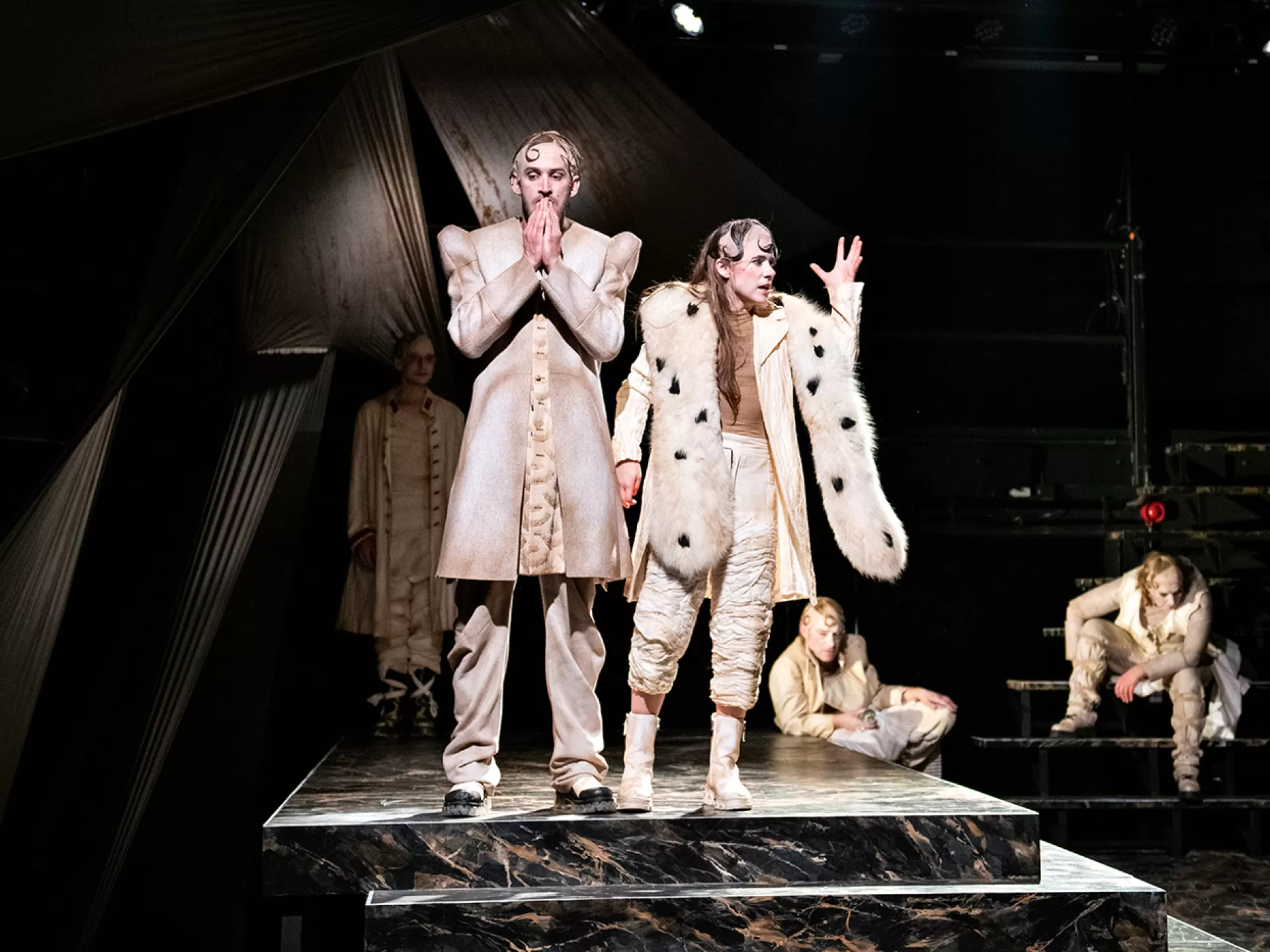 Die Figuren Friedrich (gespielt von Jonah Moritz Quast) und Elizabeth (gespielt von Jonah Moritz Quast) stehen im Vordergrund oben an einer Treppe, im Hintergrund Stallmeister, Nele und Tyll, in der Inszenierung von "Tyll" am Theater Heidelberg im Dezember 2021.