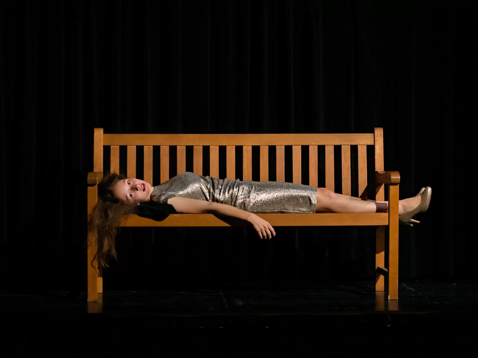 Eine Frau mit langen Haaren und in einem engen silbern glitzernden Cocktailkleid liegt auf einer Bank.