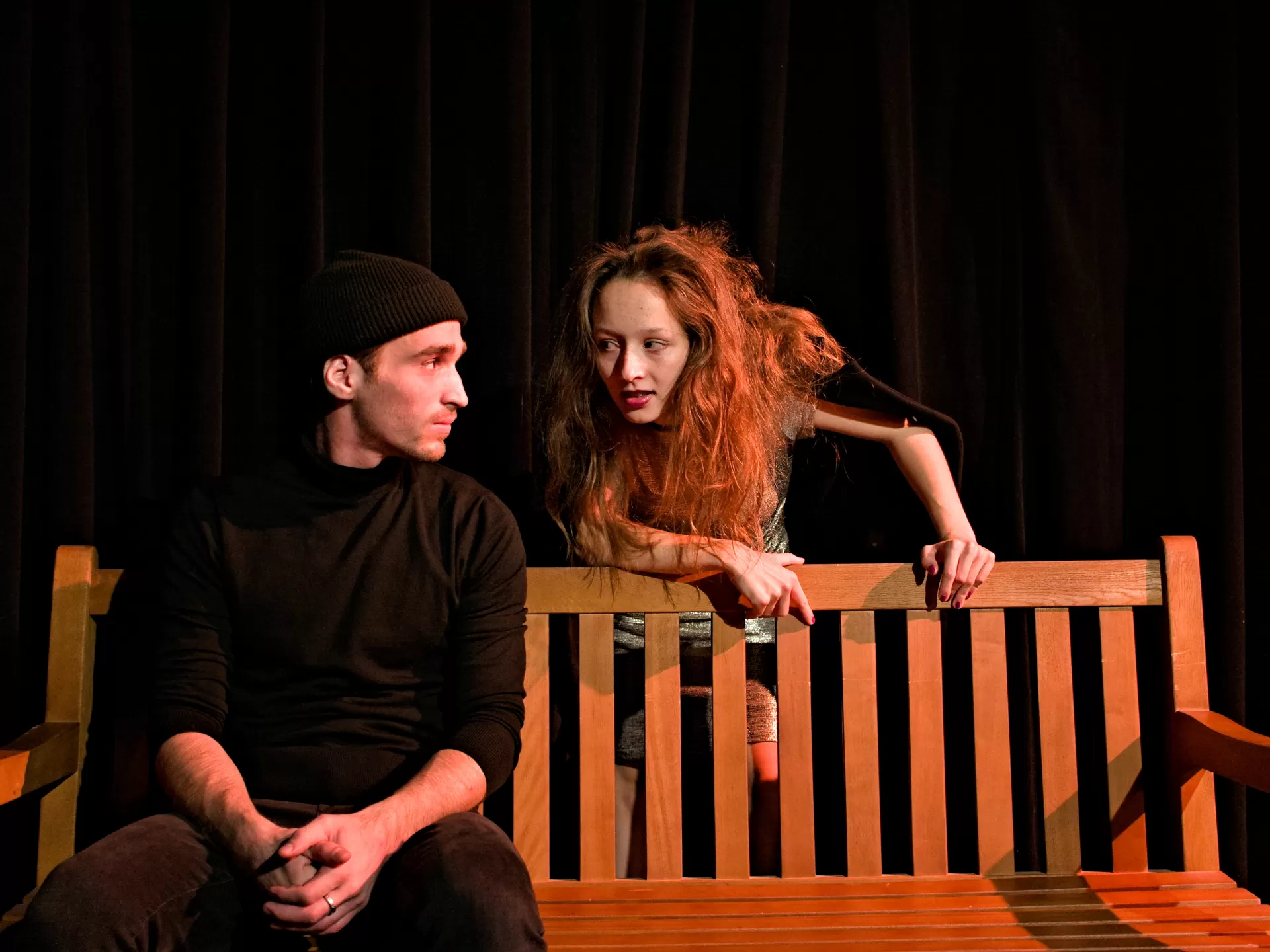 Ein Mann sitzt auf einer Bank und von hinten spricht ihn eine Frau mit langen Haaren an.