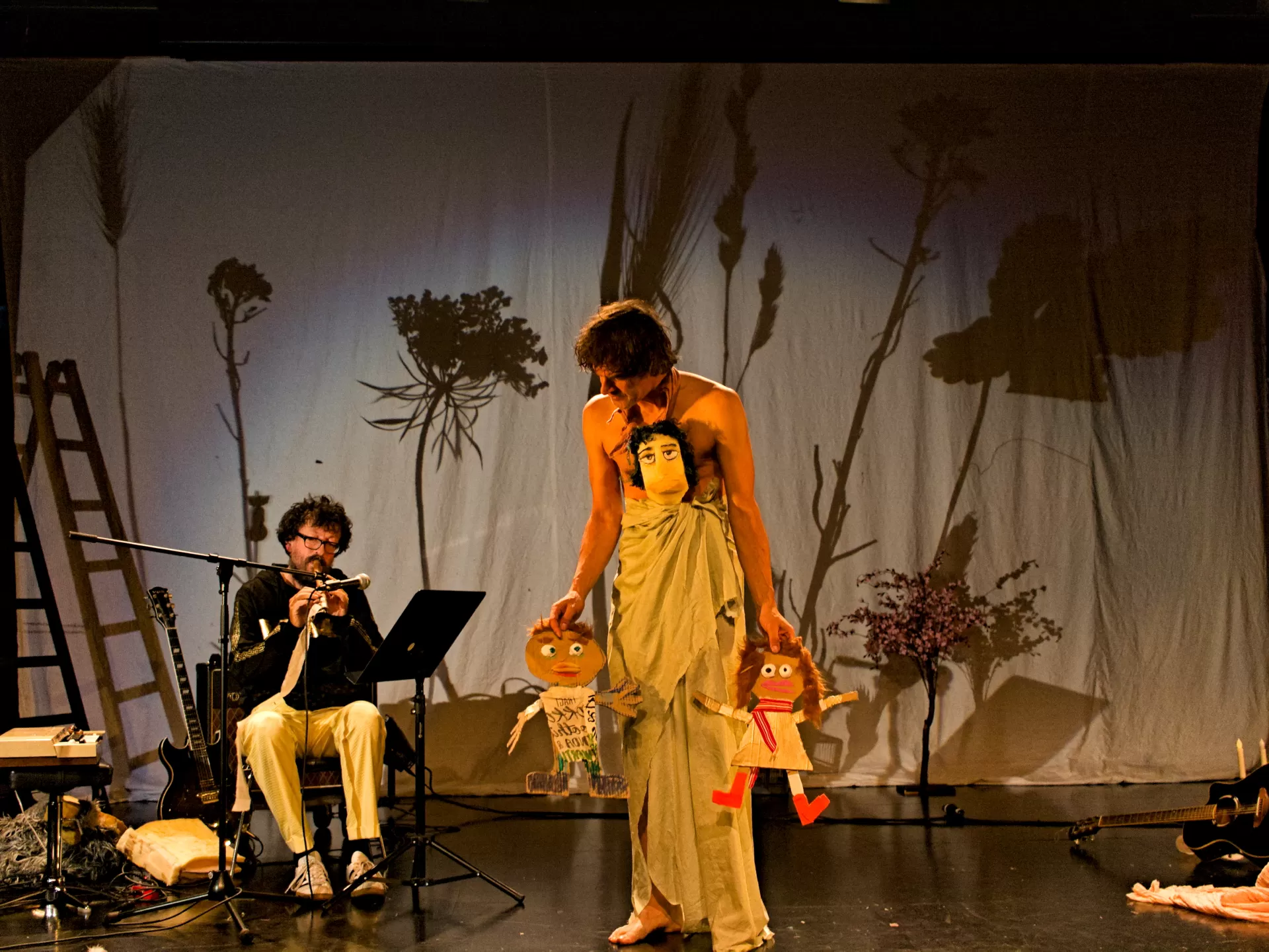 Auf der Bühne sitzt links ein Mann und spielt Flöte, in der Mitte steht ein Mann in ein Tuch gehüllt. Er hat rechts und links jeweils eine Puppe in der Hand und mittig auf seinem Brustkorb befindet sich eine Gesichtsmaske.