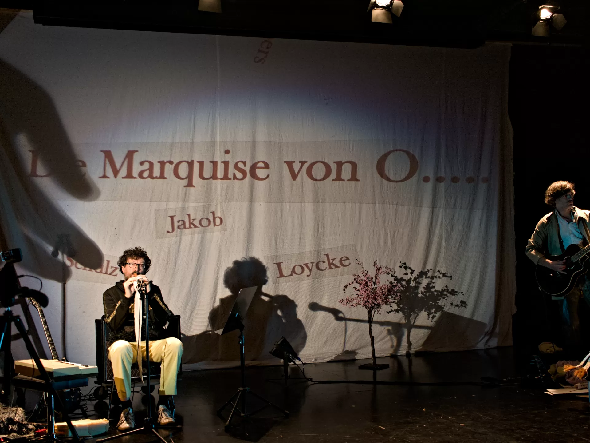 Auf der Bühne sitzt links ein Mann und spielt Flöte, rechts steht ein Mann und spielt Gitarre. Auf eine Leinwand hinter ihnen wird der Titel "Die Marquise von O...." projeziert.