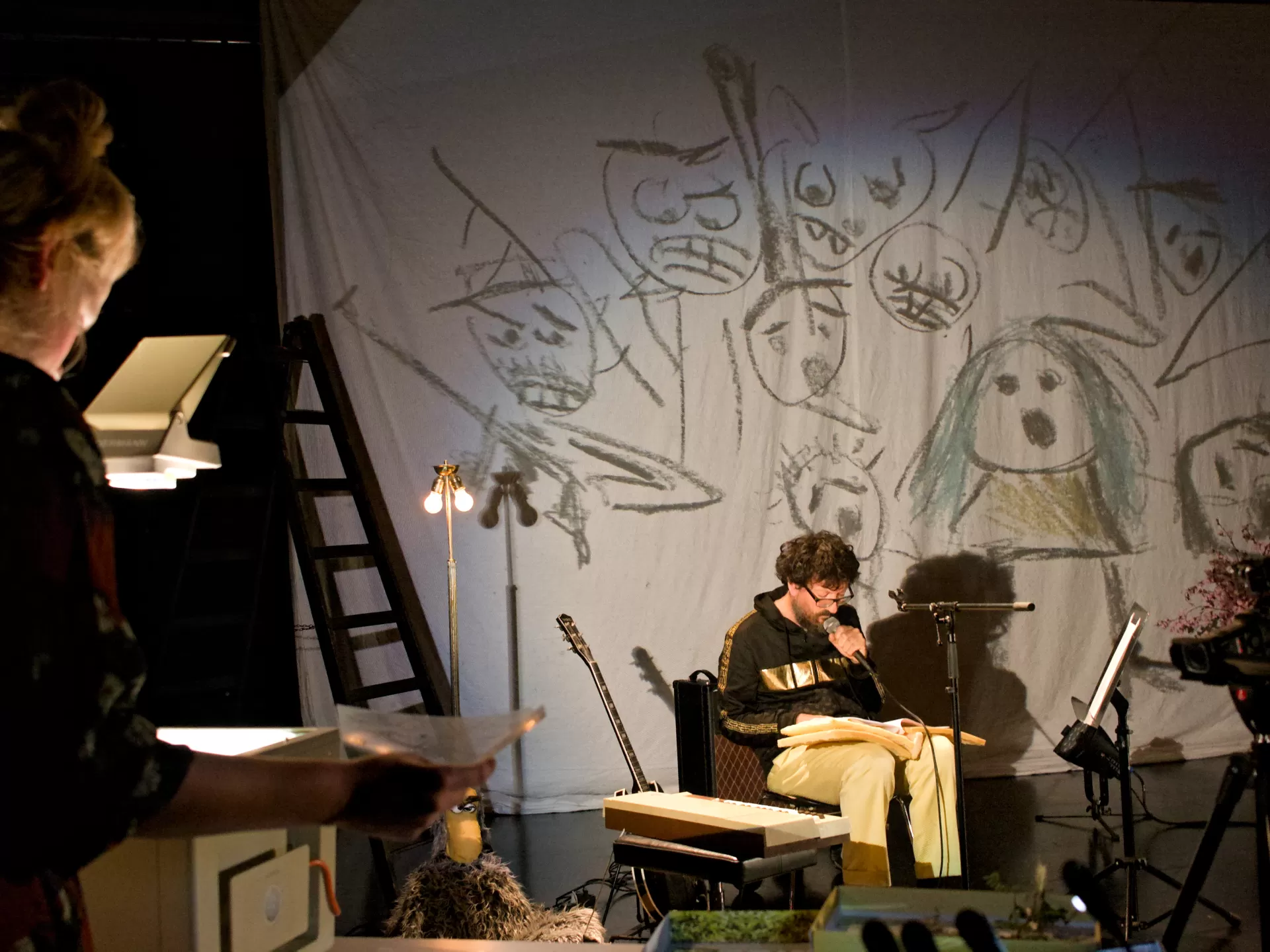 Auf der Bühne sitzt ein Mann und spricht in ein Mikrofon, neben ihm steht eine Gitarre und eine Handpuppe sitzt am Boden. Vorn links unscharf ist von hinten die Regisseurin zu sehen, die eine Projektion an die Leinwand auf der Bühne wirft. Die Projektion zeigt gekritzelte Bleistift-Gesicher.