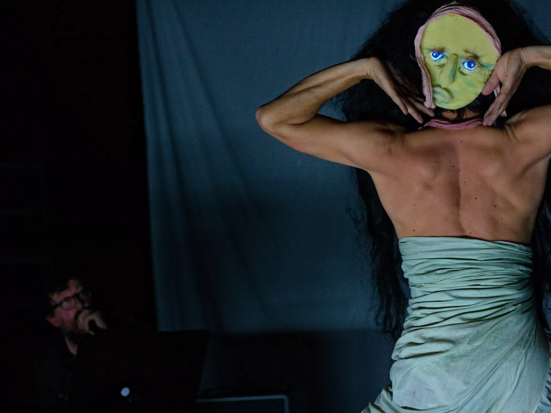 Ein Mann ist von hinten zu sehen, er hat ein blaues Tuch um den Körper gewickelt, die Schultern sind frei und auf seinem Hinterkopf ist eine gelbe Gesichtsmaske zu sehen.