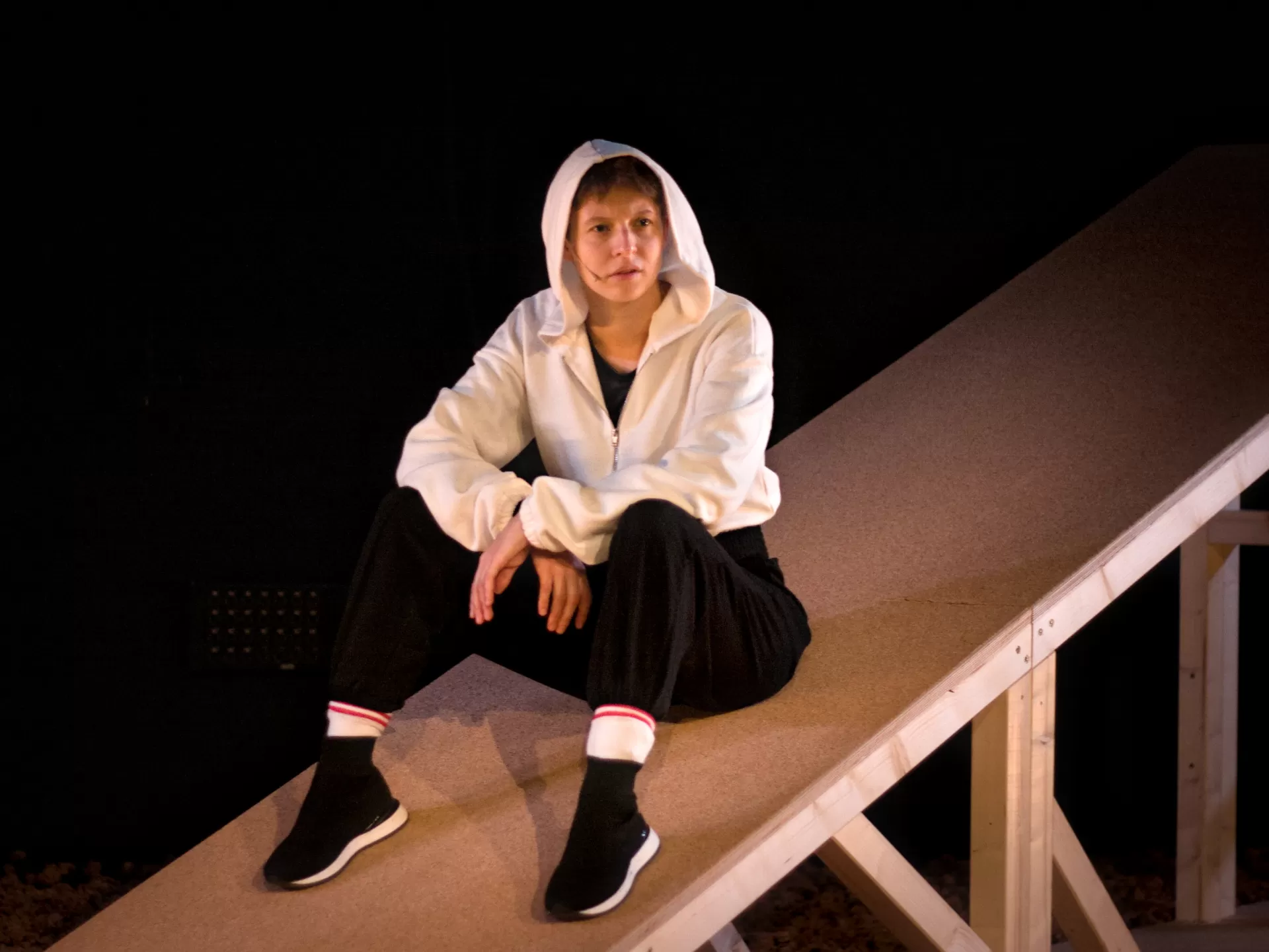Eine Frau sitzt auf einer Schräge aus Holz und schaut nachdenklich.