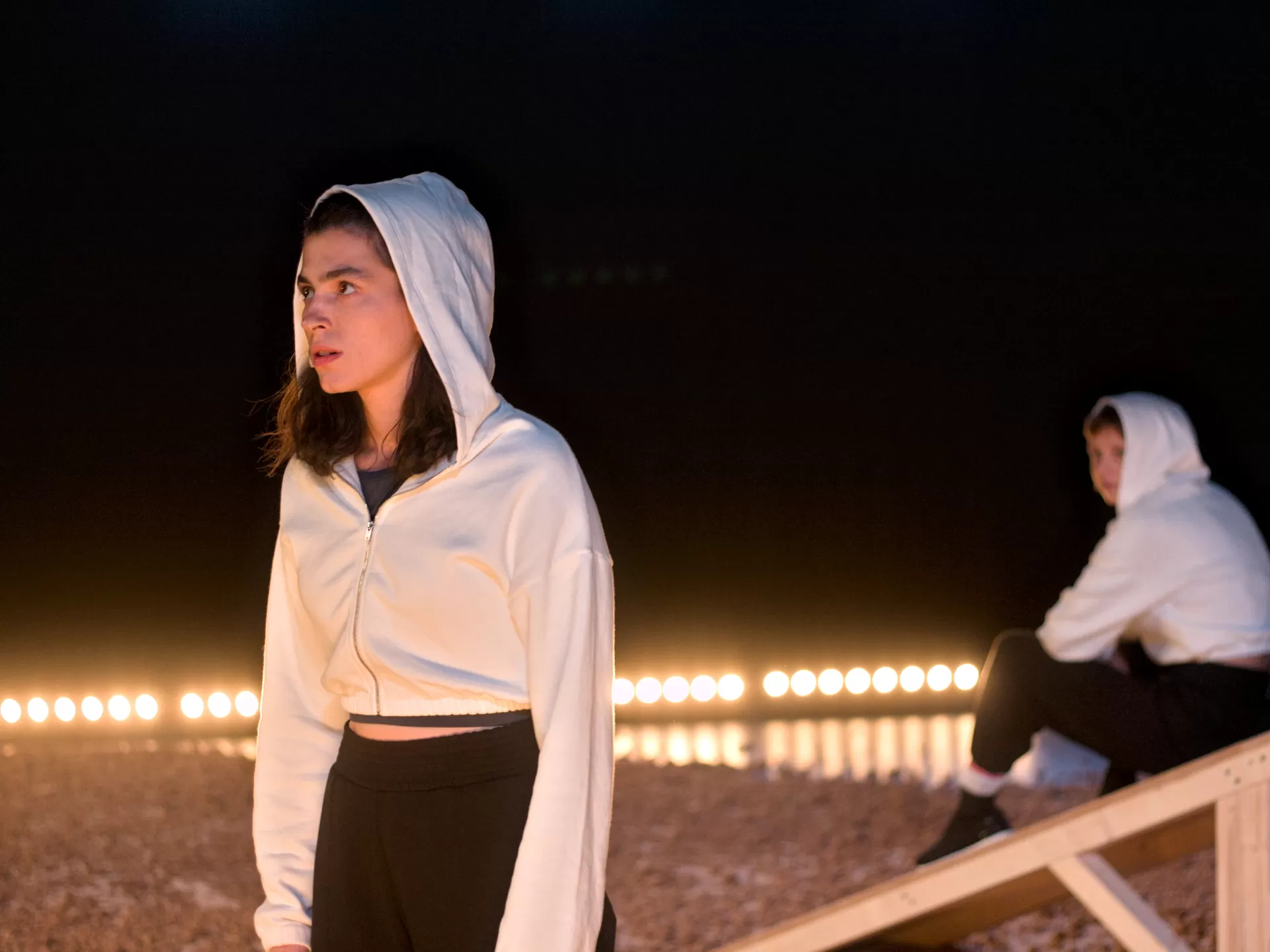 Eine Frau im Vordergrund blickt ernst, im unscharfen Hintergrund sitzt rechts eine zweite Frau auf einer Holzschräge.