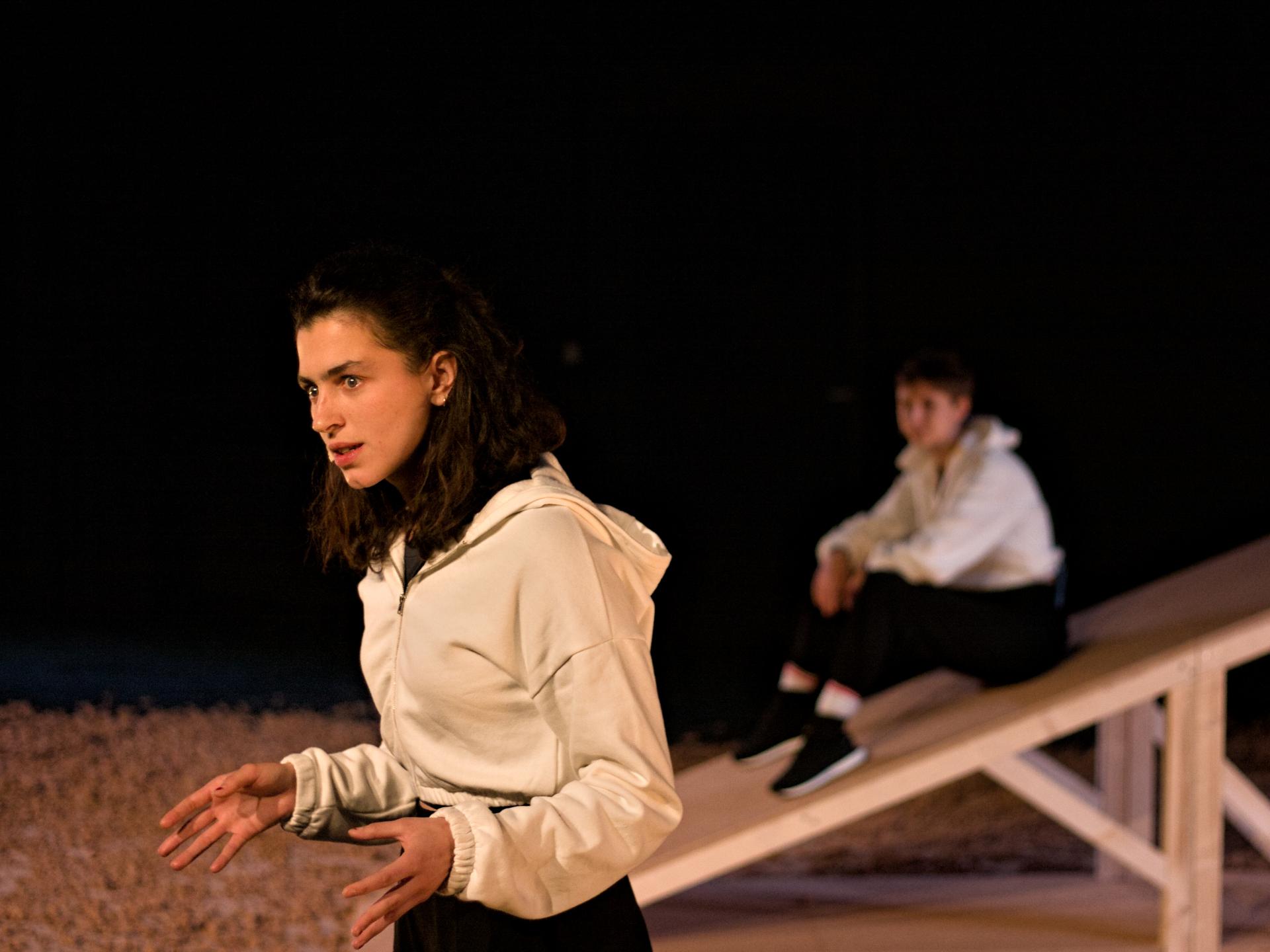 Eine Frau im Vordergrund steht mit beiden Händen vor dem Körper und spricht, im Hintergrund sitzt eine zweite Frau auf einer Holzschräge.