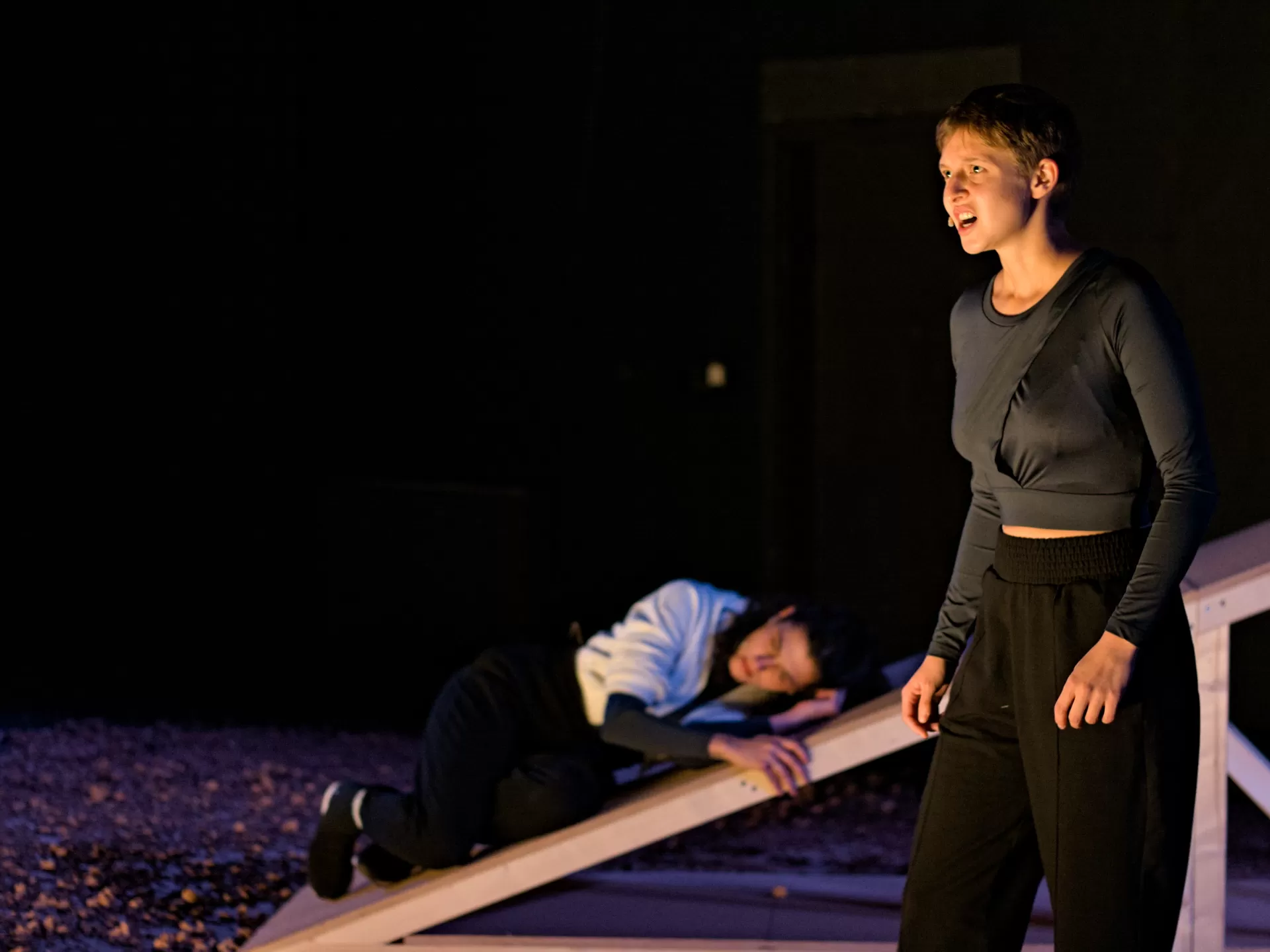 Eine Frau liegt gekauert auf einer Schräge aus Holz, eine zweite Frau steht davor und spricht.