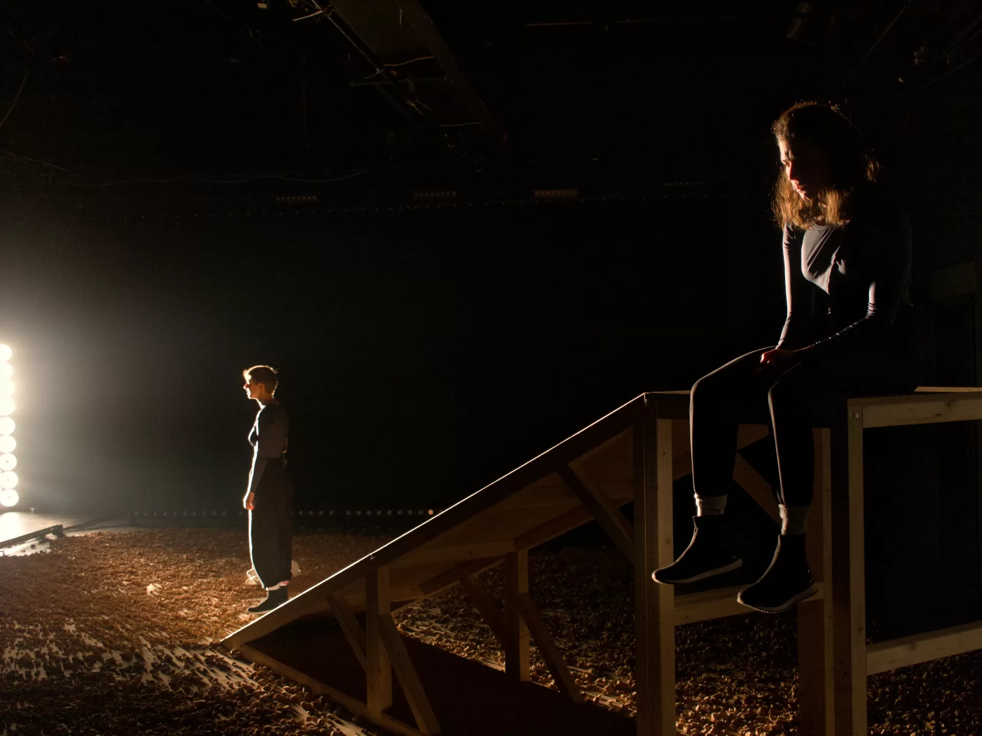 Eine Frau  steht vor hellem Scheinwerferlicht, rechts daneben sitzt eine zweite Frau auf der Kante auf einem Holzpodest und blickt nachdenklich nach unten.