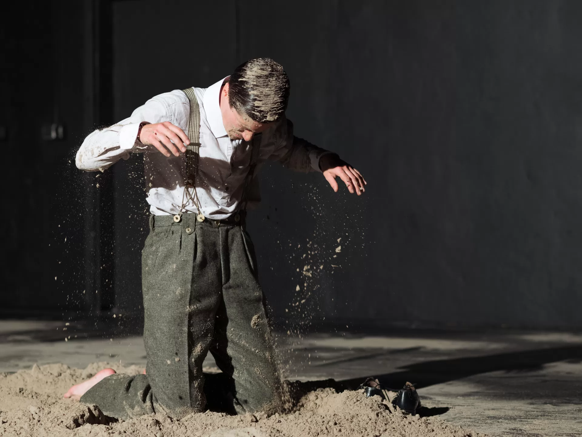 Mann kniet mit geschlossenen Augen in Sand auf Bühne, hat Sand im Haar.