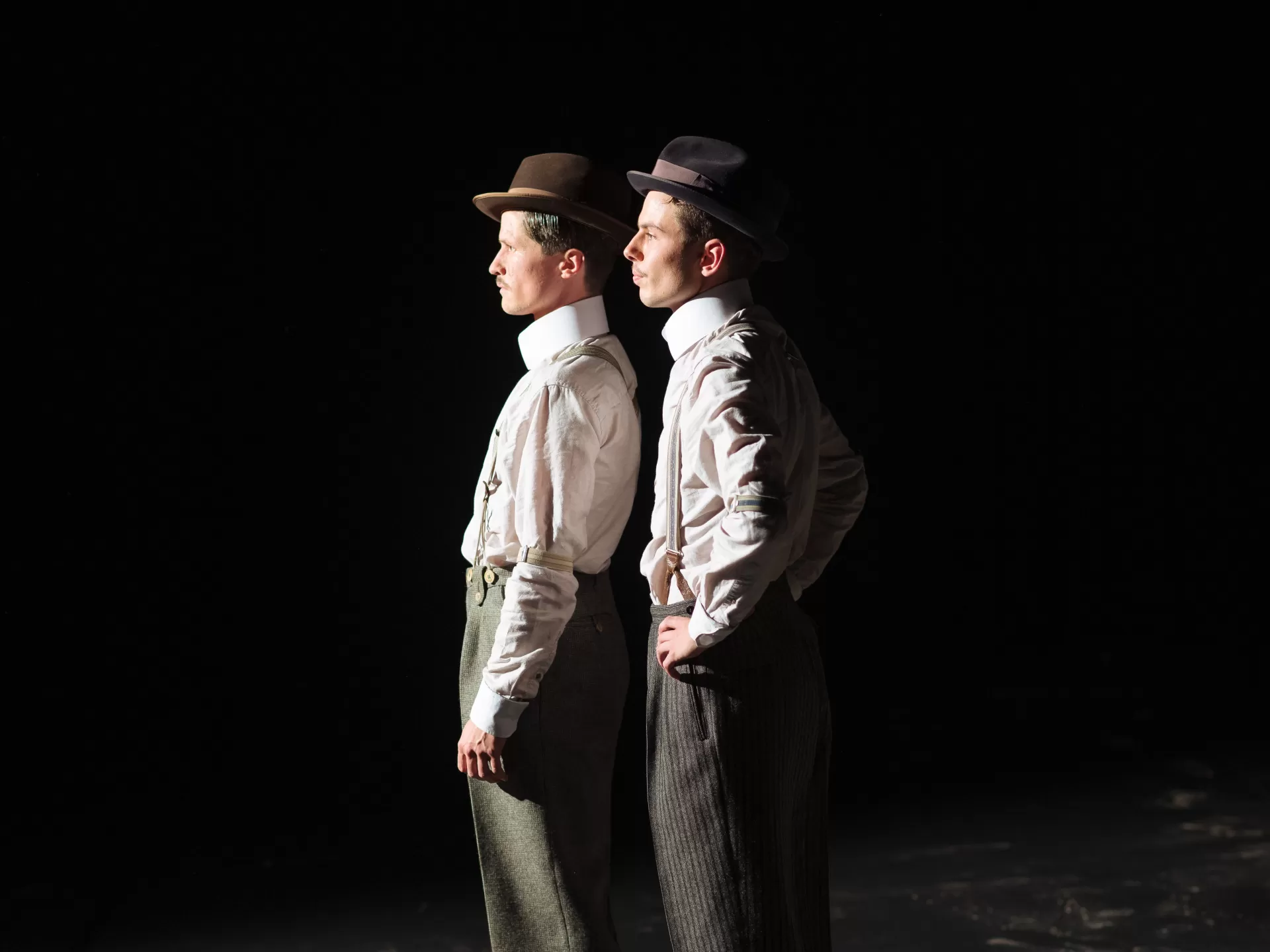 Blick von der Seite auf zwei Männer, die hintereinander, beide nach links schauend, vor einem schwarzen Hintergrund stehen. Jeder hat einen Hut auf, ein weisses Hemd und eine Hose mit Hosenträgern.