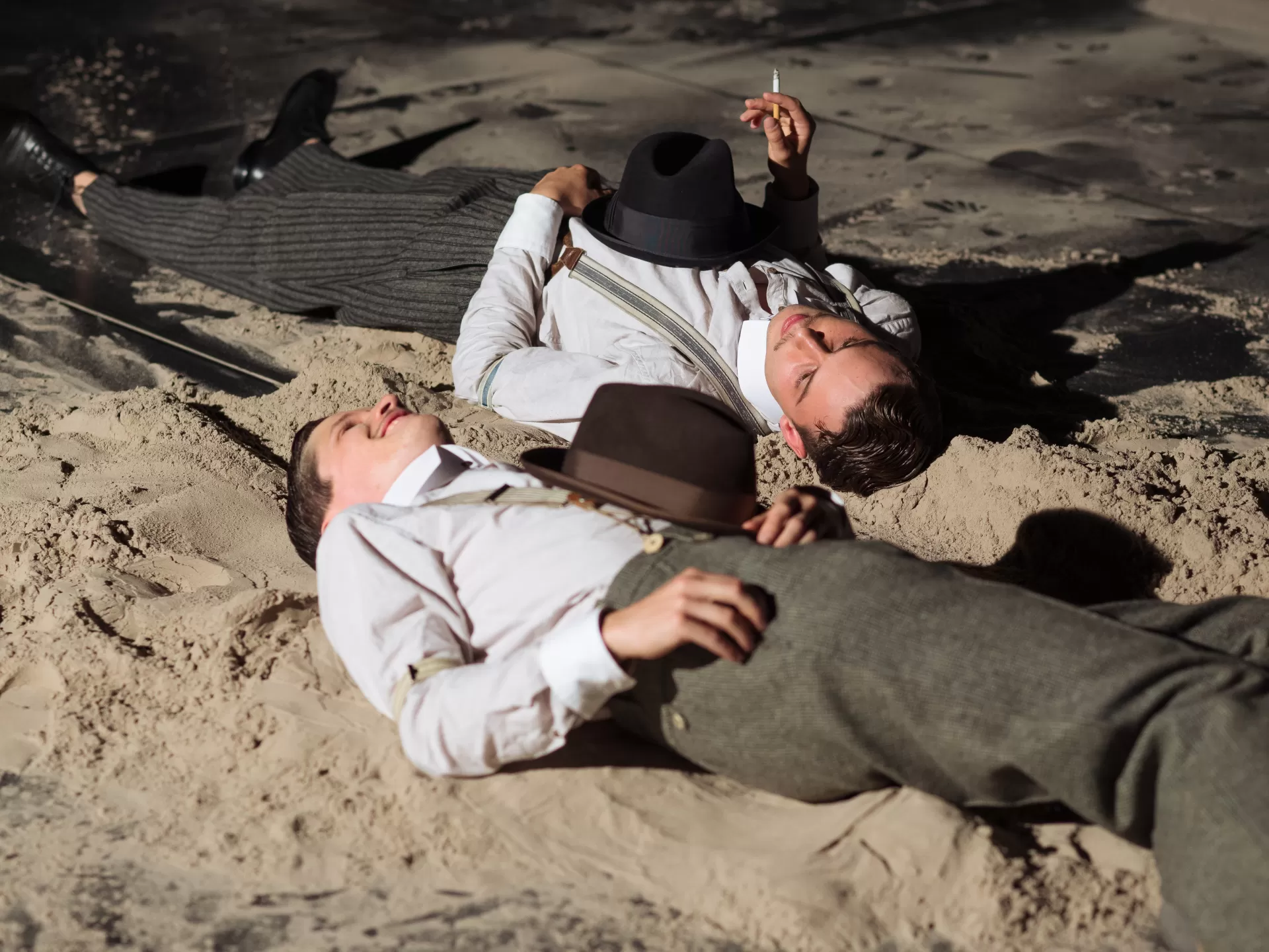 Blick von schräg seitlich auf zwei Männer am Boden liegende Männer, in Sand auf einer Bühne. Jeder hat ein weisses Hemd und eine Hose mit Hosenträgern an. Beide haben einen Hut auf dem Brustkorb liegen. Der hintere der zwei hat eine Zigarette in der Hand..