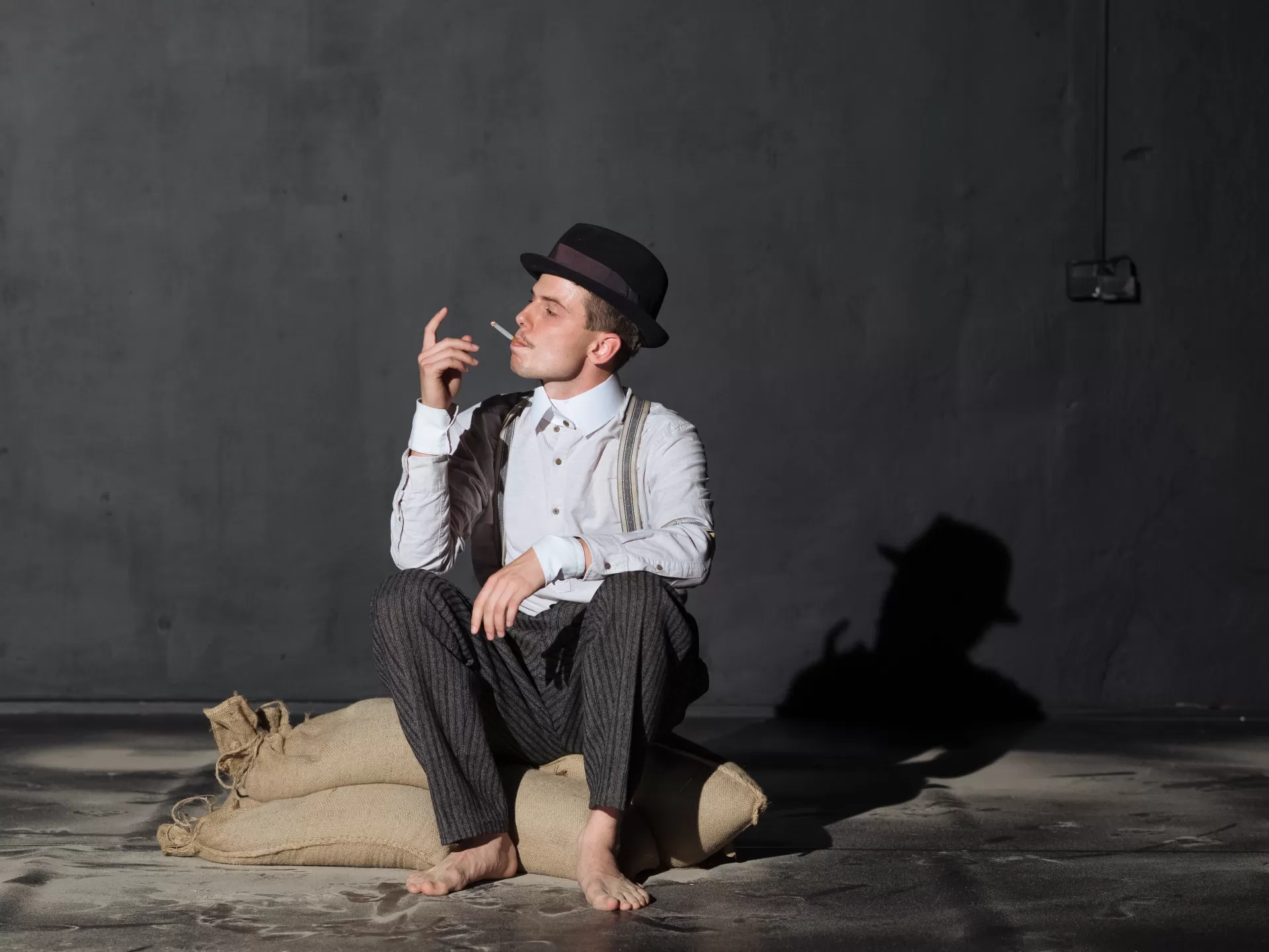 Ein Mann sitzt auf zwei flachen am Boden liegenden Leinensäcken auf einer Bühne, um ihn herum ist Sand verstreut. Der Mann trägt ein weisses Hemd, eine Hose mit Hosenträgern und einen Hut. Er ist barfuß und hat eine Zigarette im Mund. Hut auf dem Brustkorb liegen. Der hintere der zwei hat eine Zigarette in der Hand..