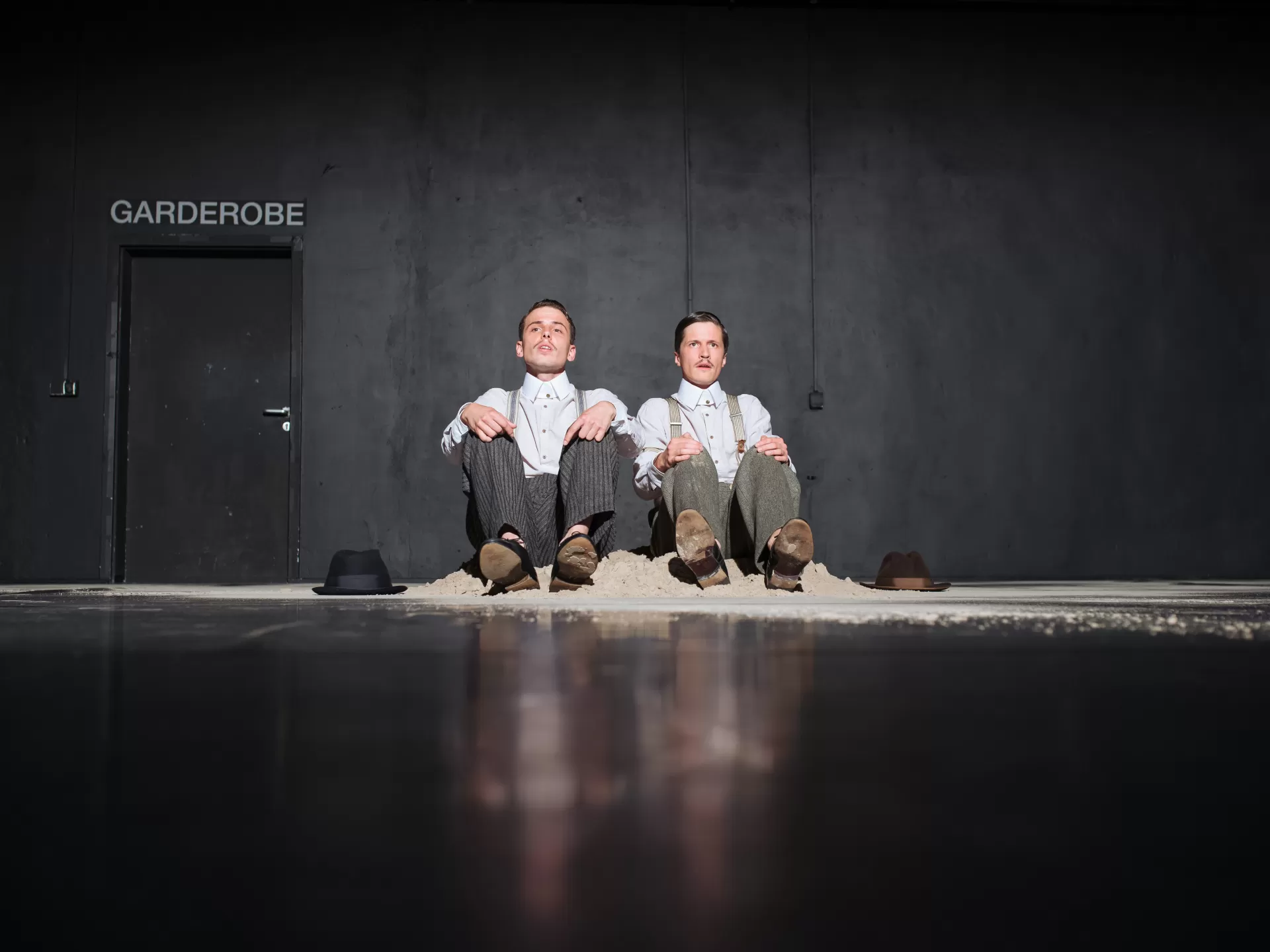 Zwei Männer sitzen nebeneinander am Boden in einem kleinen Sandhaufen vor einer schwarzen Wand. Jeder hat ein weisses Hemd und eine Hose mit Hosenträgern an, neben ihnen liegt jeweils ein Hut am Boden..