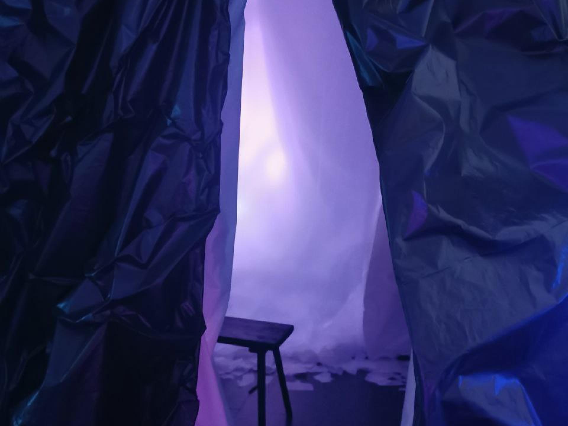 Zu sehen ist das Ende und zwei Beine einer hölzernen Sitzbank in einem Raum mit blau-lila Licht, umgeben von dunklen, lilafarbenen Vorhängen aus Papier oder Stoff.