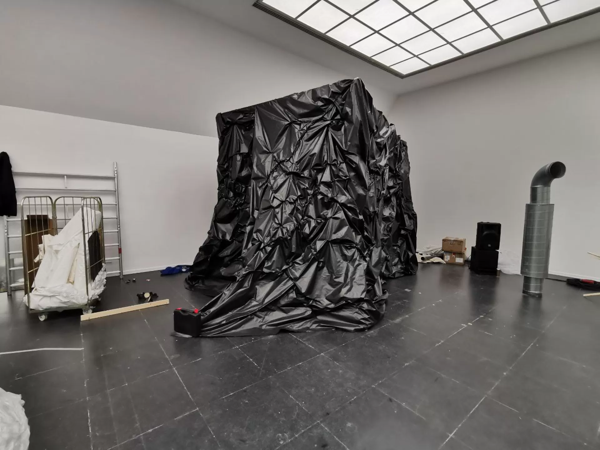 Zu sehen ist ein großer schwarzer Quader, höher als ein Mensch, eingepackt in schwarze Folie. An der Vorderseite steht auf einem Zipfel der Folie ein schwarzer Plastikkanister. Im Raum um den Quader herum stehen wie zufällig angeordnet zwei Packkisten, ein mannshohes metallenes Rohr und ein Gitterwagen mit weissen Stoffrollen.