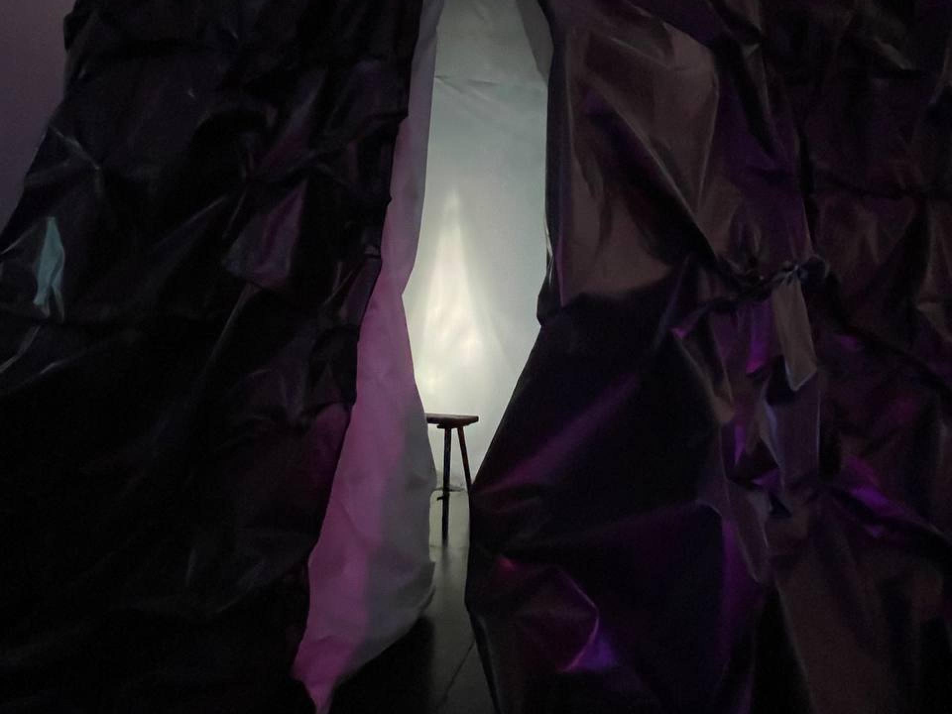 Zu sehen ist das Ende und zwei Beine einer hölzernen Sitzbank in einem Raum mit weissem Licht, umgeben von dunklen, lilafarbenen Vorhängen aus Papier oder Stoff.