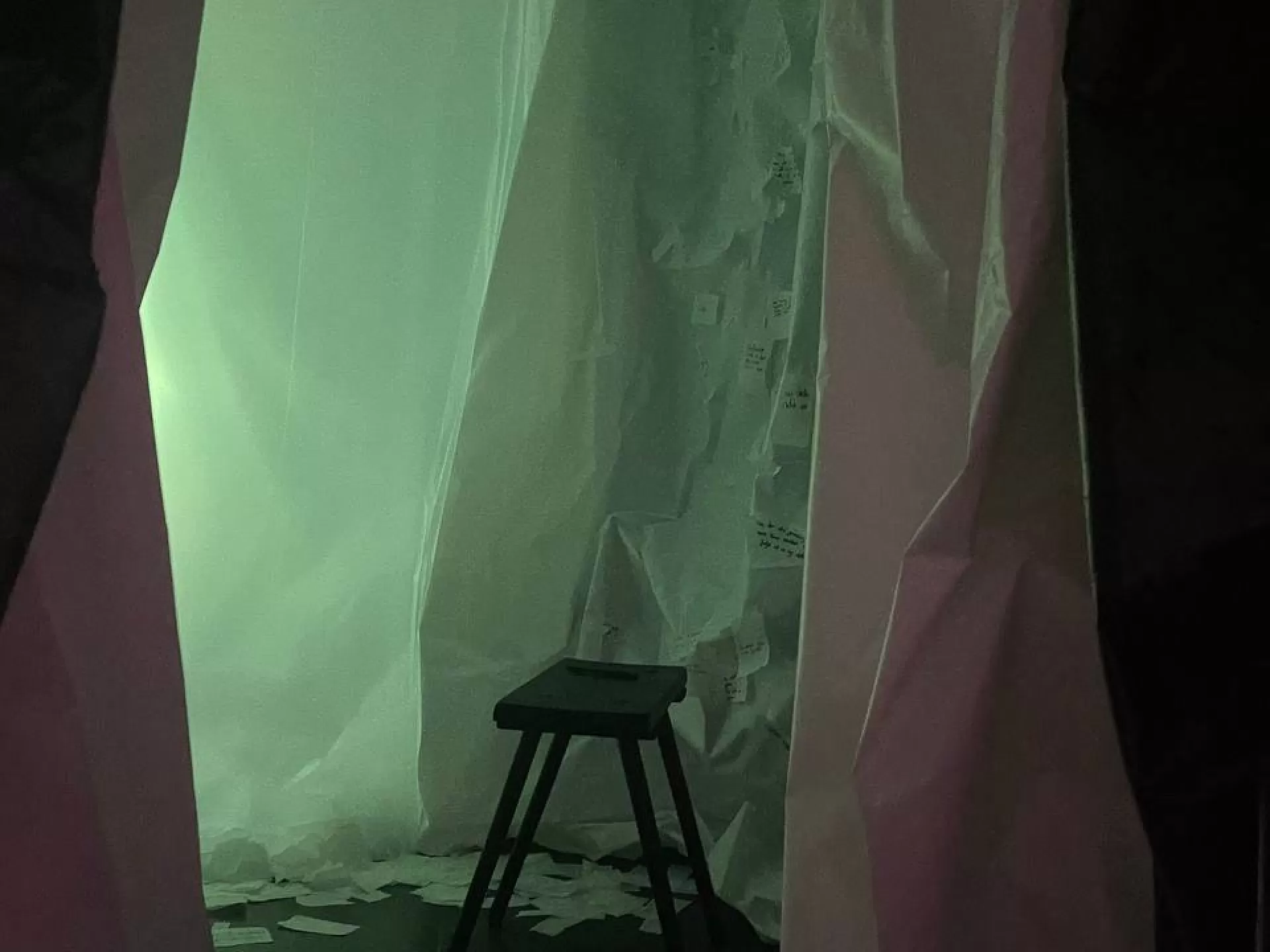 Zu sehen ist eine hölzerne Sitzbank mit vier Beinen in einem Raum mit grünemLicht, umgeben von dunklen Vorhängen aus Papier oder Stoff. Am Boden liegen durcheinander weisse Papierblätter, an der Wand hängen verschiedene Papierfetzen mit Schrift darauf.