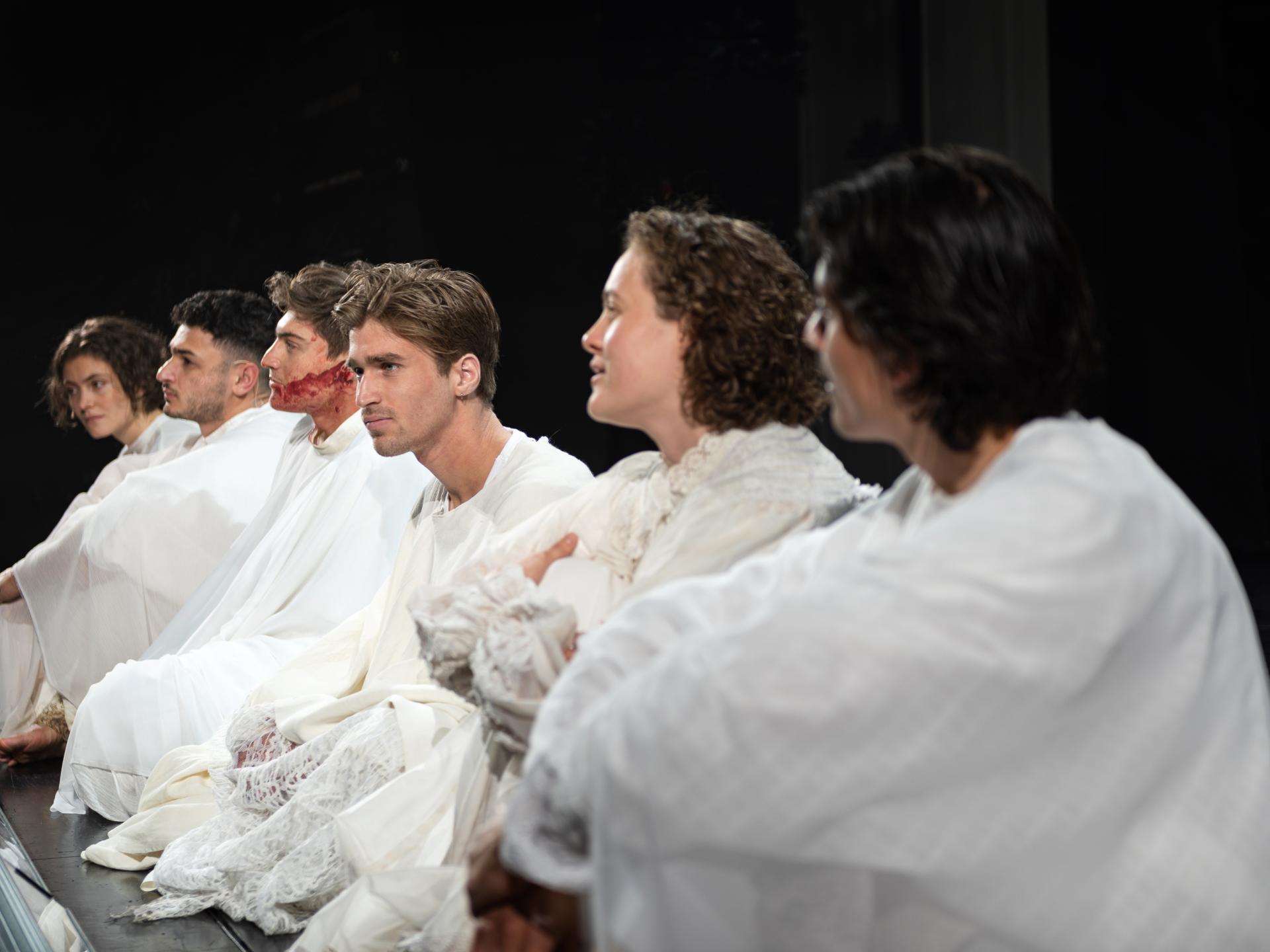 Ensemble, alle in Weiß gekleidet, sitzt vorne an der Bühnenkante auf dem Boden.