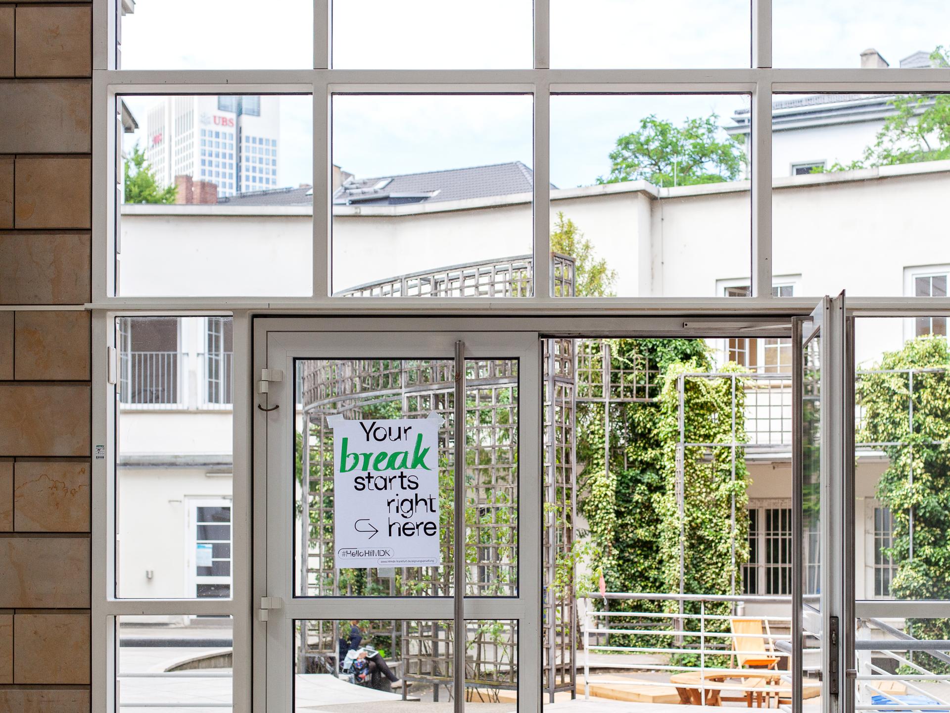 Offene Glastür zum Innenhof der HfMDK mit einem Plakat auf dem steht: Your Break starts right here.