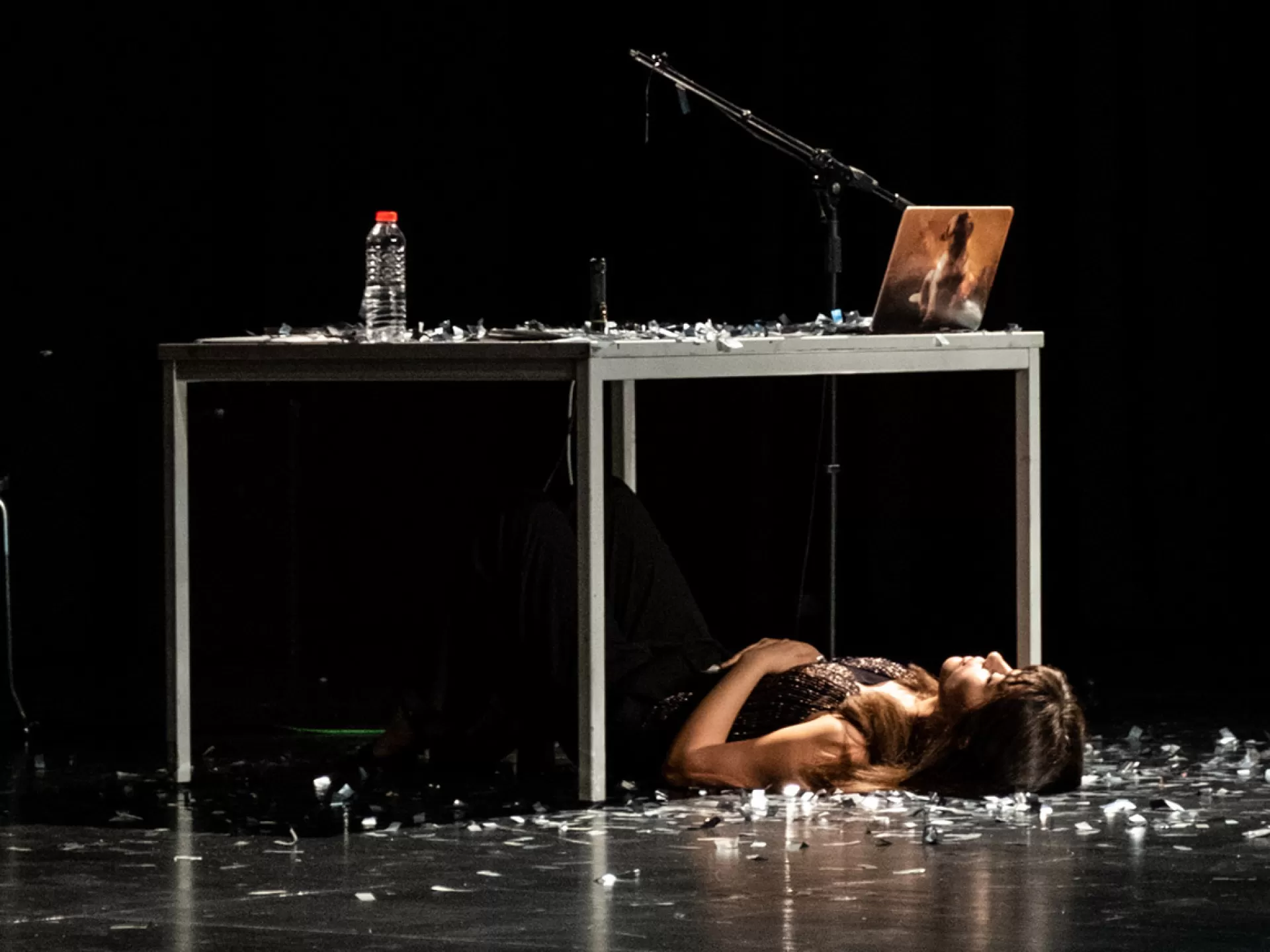 Eine Frau liegt am Boden auf dem Rücken unter einem Tisch, auf dem Boden liegt silberner Flitter. Auf dem Tisch über ihr stehen eine Wasserflasche und ein Laptop.