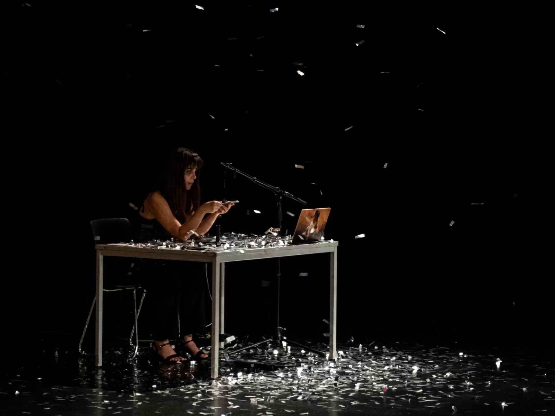 Dunkler Bühnenraum, eine junge Frau sitzt an einem Tisch, sie hält ein Handy in der Hand und blickt darauf.