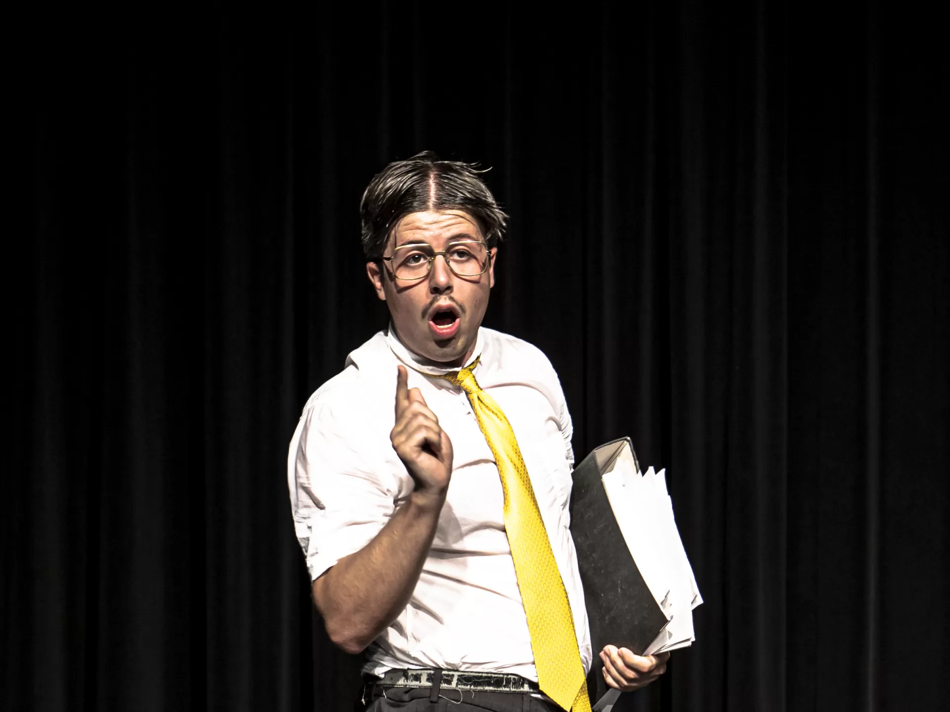 Schauspieler mit Mittelscheitel, Brille, Ordner, gelber Krawatte und erhobenem Zeigefinger.