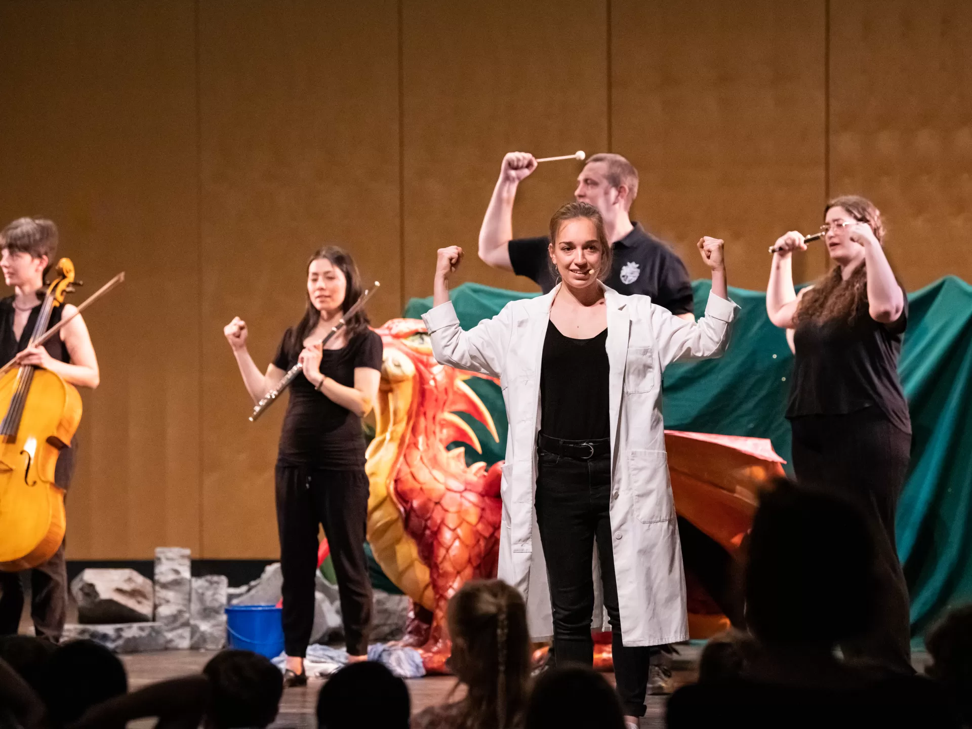 Künstlerinnen auf der Bühne bei einem Kinderkonzert. Im Vordergrund eine junge Frau im weißen Kittel, die ihre Arme hebt.