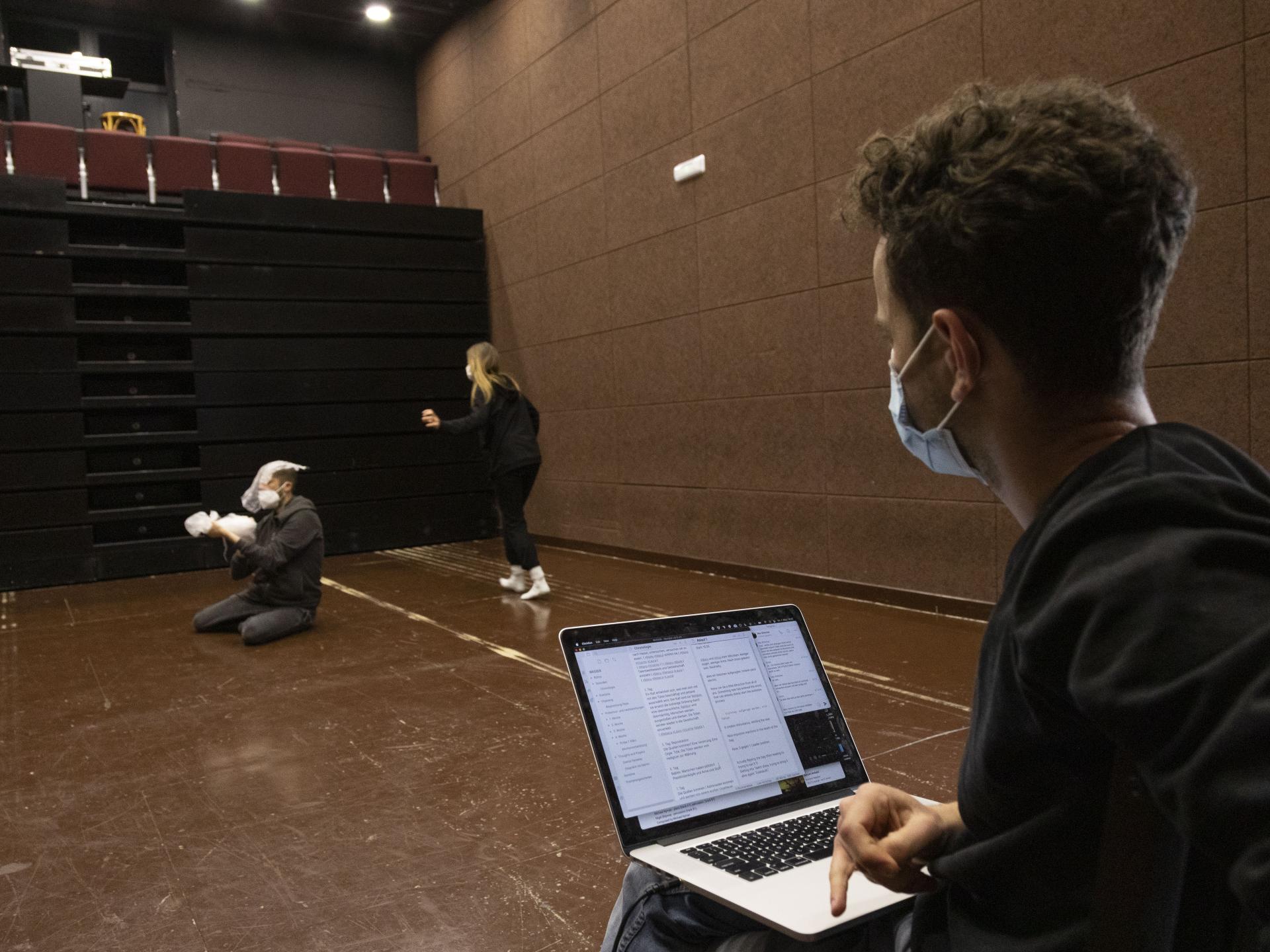 Mann sitzt mit Laptop auf den Knien, darauf sind Texte zu sehen, er beobachtet zwei Spieler*innen im Raum bei Proben.