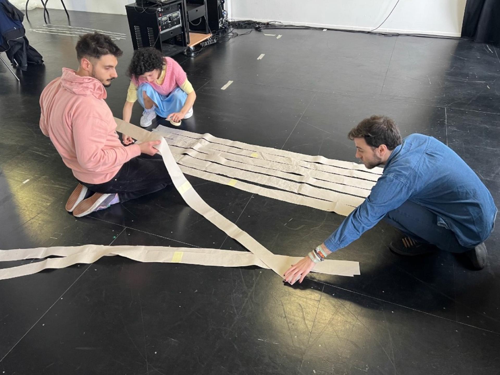 Drei Studierende, eine Frau und zwei Männer, knien am Boden und legen ca. ein Meter lange und 10 cm breite Papierstreifen nebeneinander.