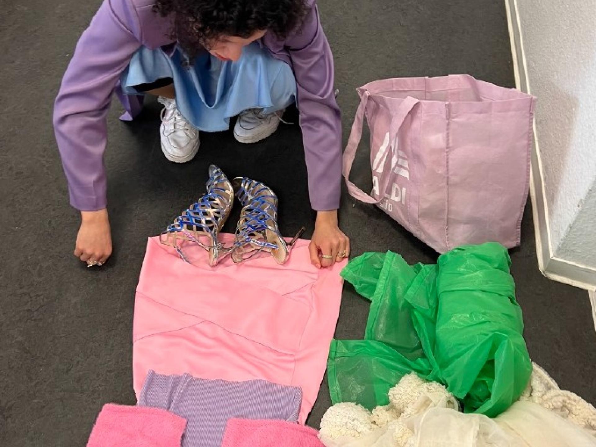 Eine Frau kniet vor und betrachtet auf dem Boden liegende Kleidungsstücke und Stoffe: eine rose Jacke, ein rosa Rock, grüne und weisse Stoffe.