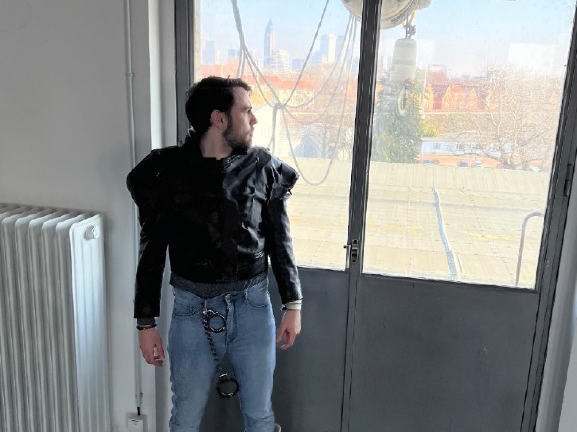 Ein Mann gekleidet in Jeans, einer kurzen schwarzen Lederjacke und weissen Gummistiefeln steht vor einem Fenster.
