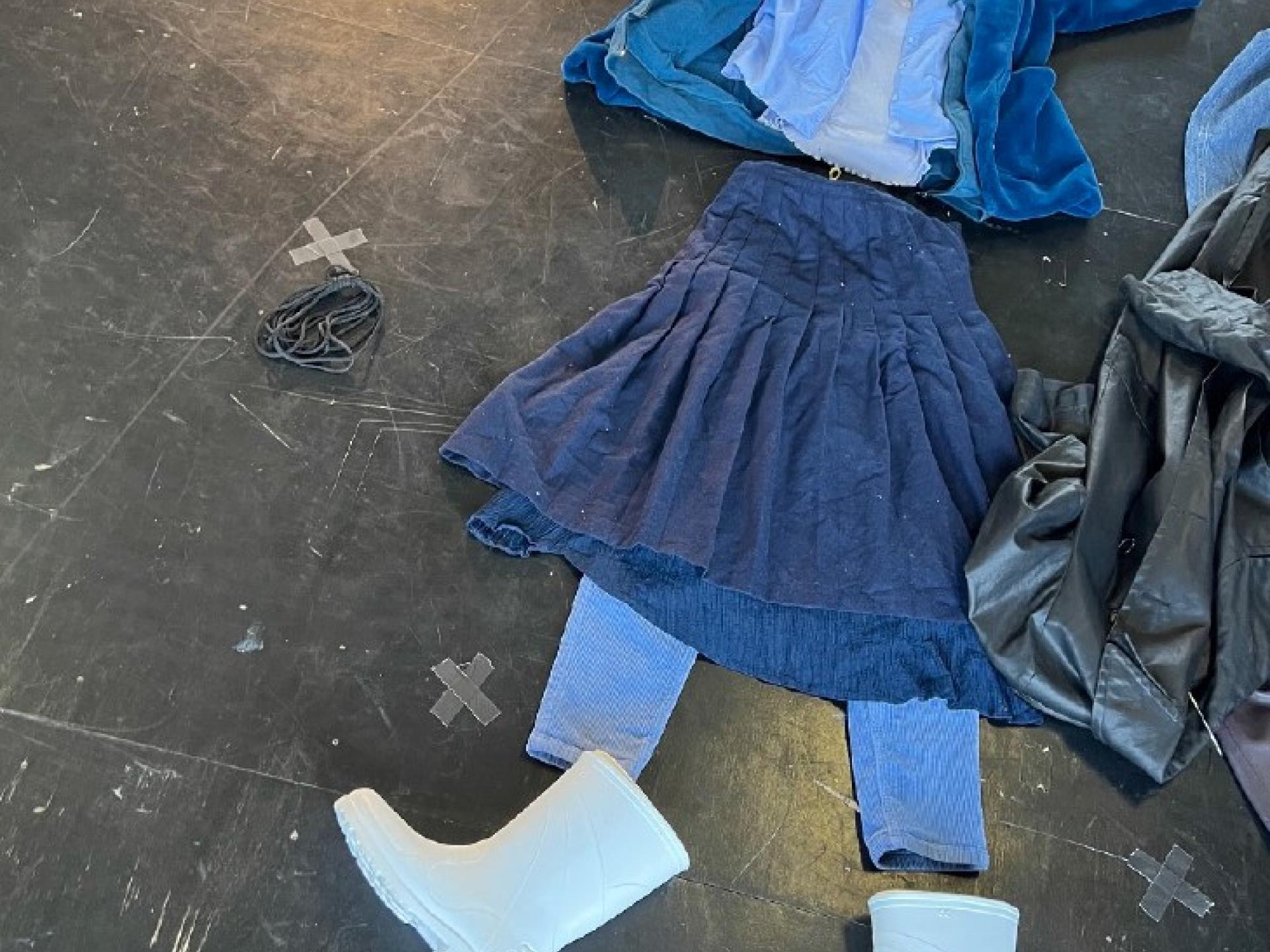 Auf dem Boden liegend Kleidungsstücke in verschiedenen Blautönen: ein Rock, eine Hose, ein Hemd, eine Jacke und weisse Gummistiefel.