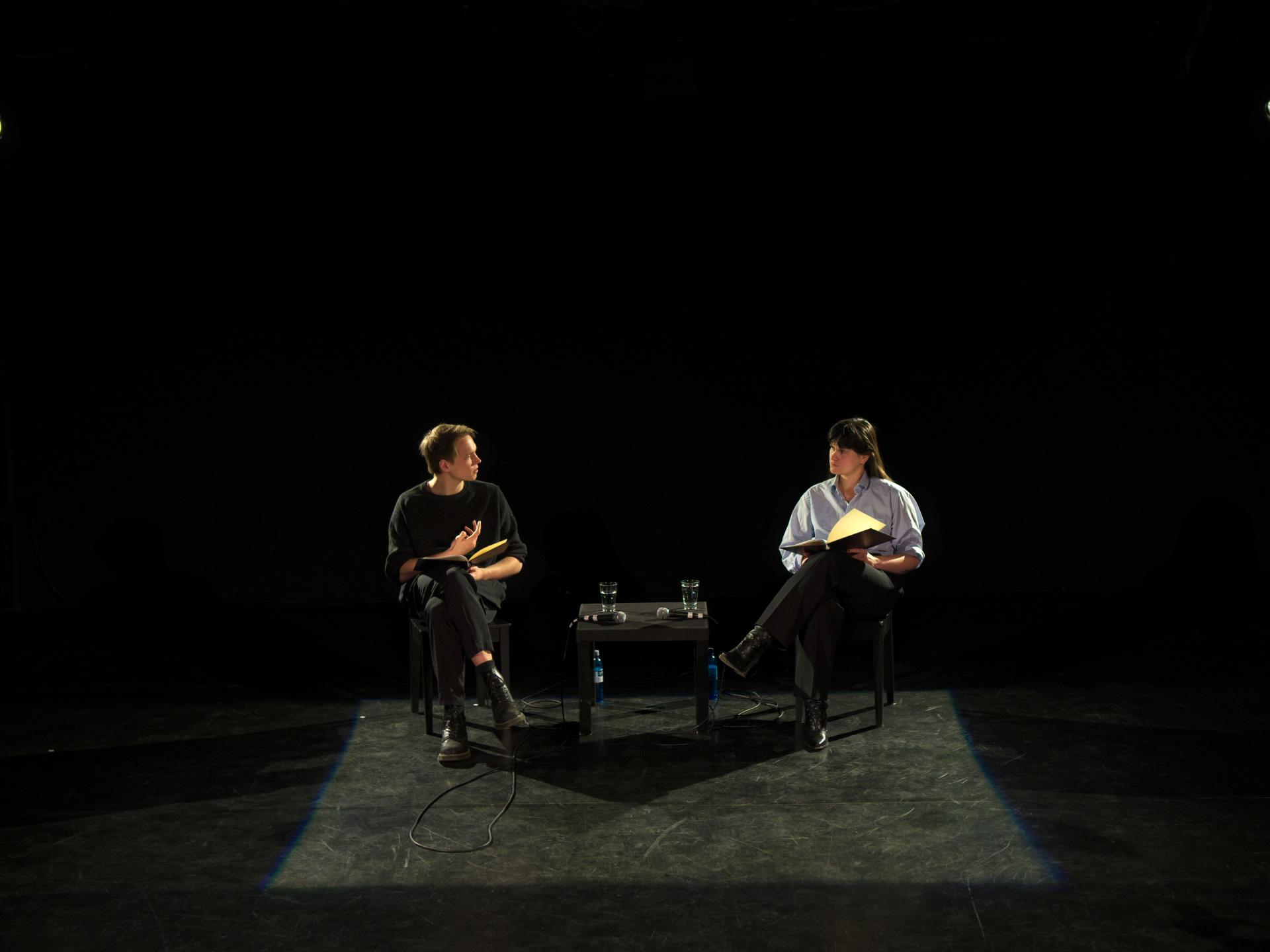 Ein Schauspieler und eine Schauspielerin sitzen in einem dunklen Raum auf Stühlen und sprechen zueinander 