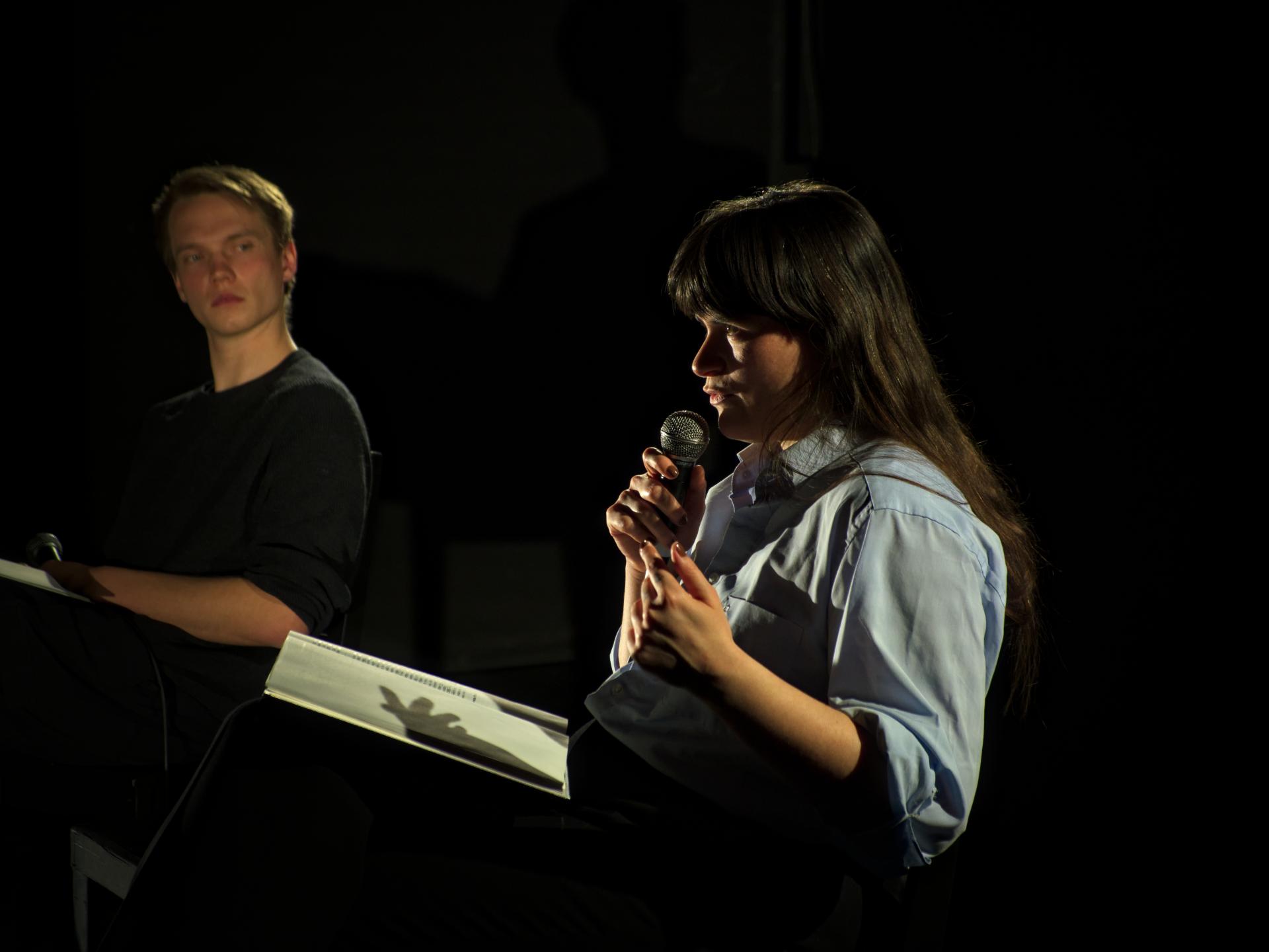 Eine Schauspielerin spricht in ein Mikrofon, im Hintergrund schaut ihr ein Schauspieler aufmerksam zu