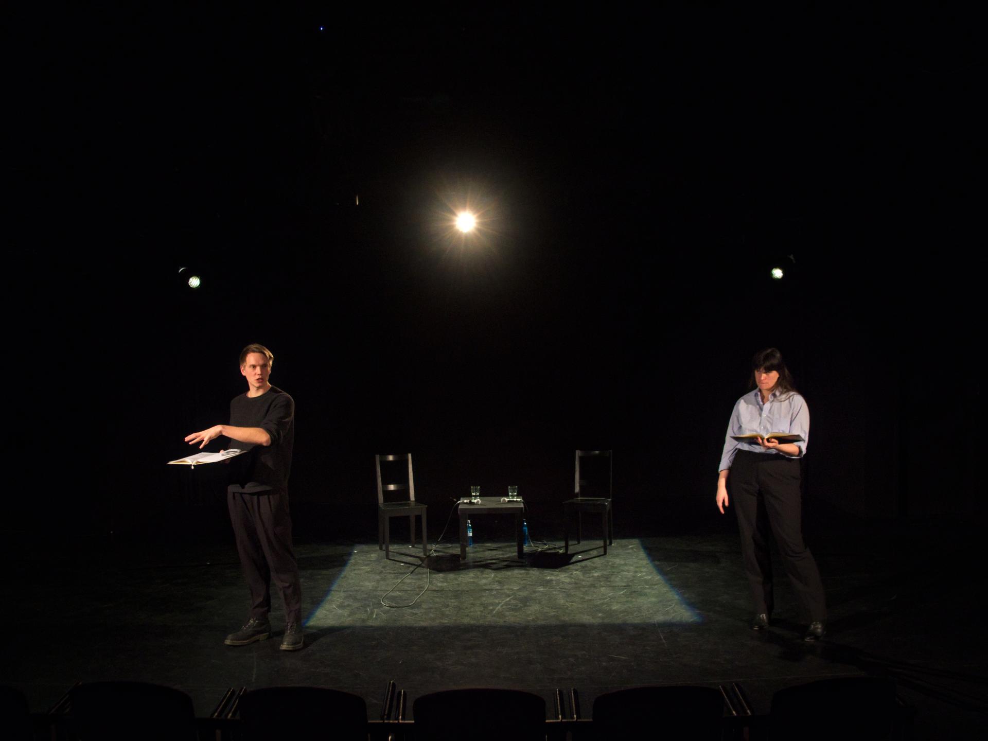 Ein Schauspieler und eine Schauspielerin stehen neben einem Licht-Quadrat, in dem Stühle und ein Tisch sind. Sie scheinen etwas zu erklären