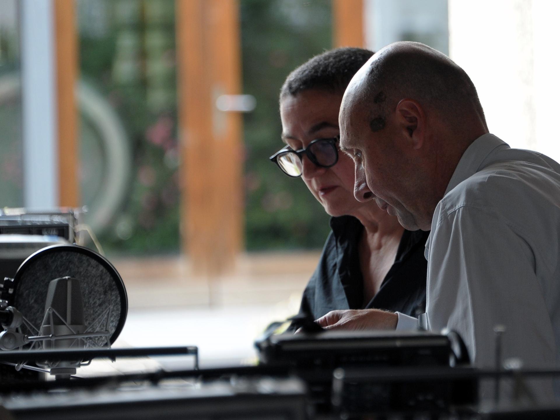 Annette Schmucki und Reto Friedmann sitzen an einem Tisch mit Mikrofonen und anderen technischen Geräten.