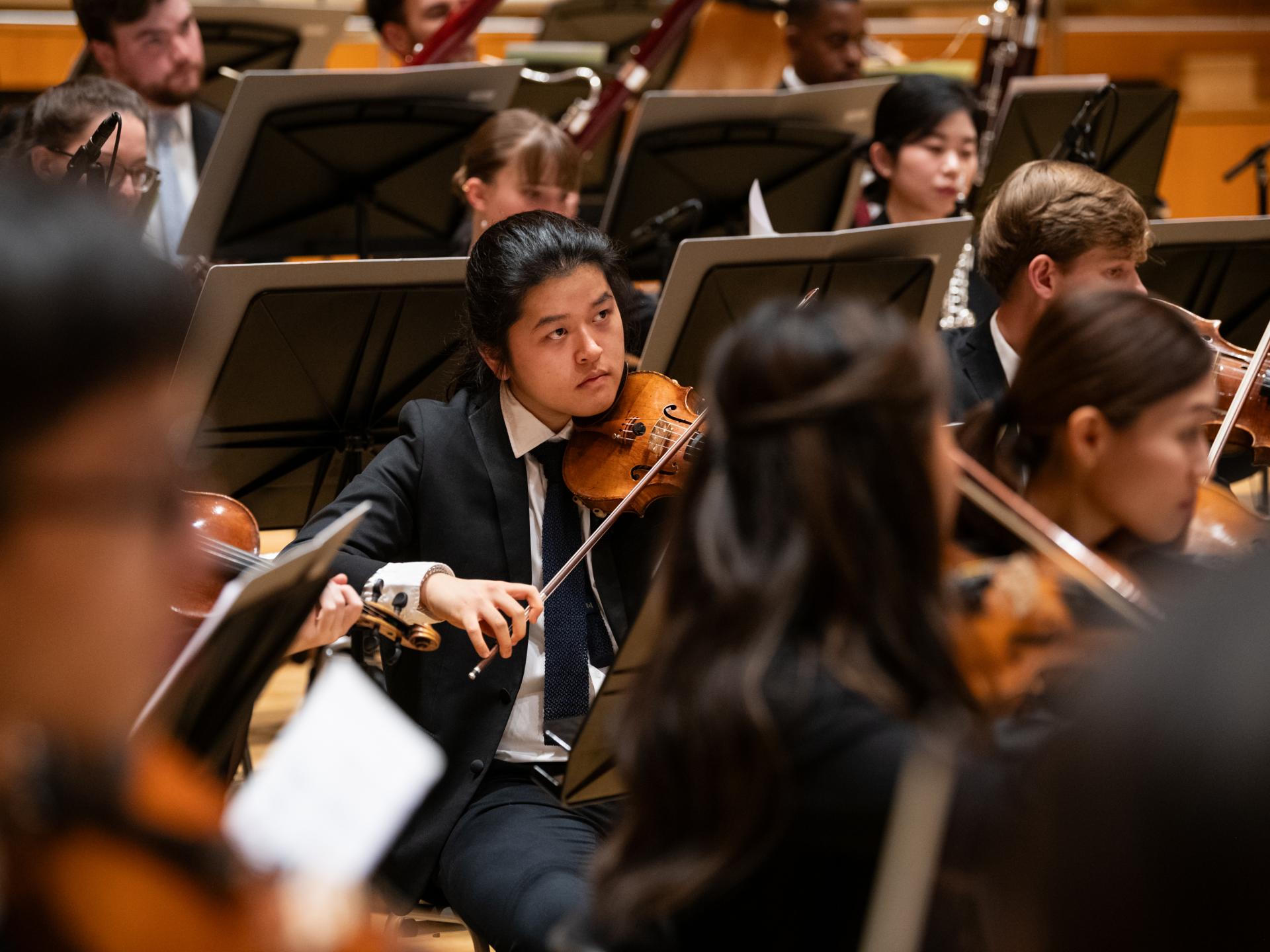 Ein Student inmitten des Orchesters spielt Geige und schaut zum Dirigenten