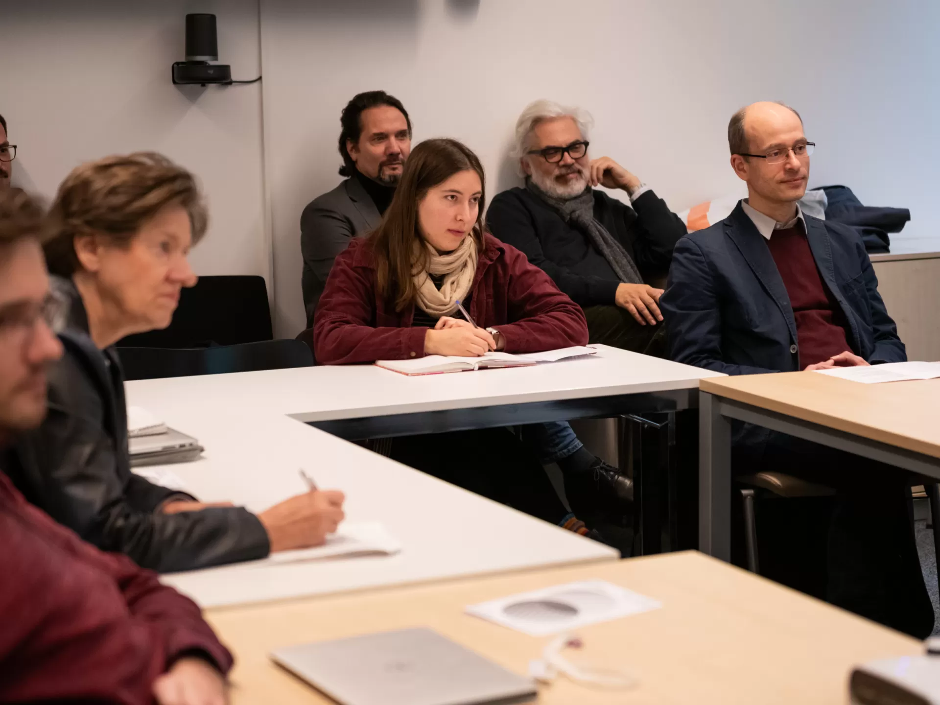 Eine Gruppe von sieben Zuhörenden sitzt in einem Vorlesungssaal an Tischen. Sie richten die Aufmerksamkeit nach rechts vorne, aber die präsentierende Person selbst ist nicht im Bild.