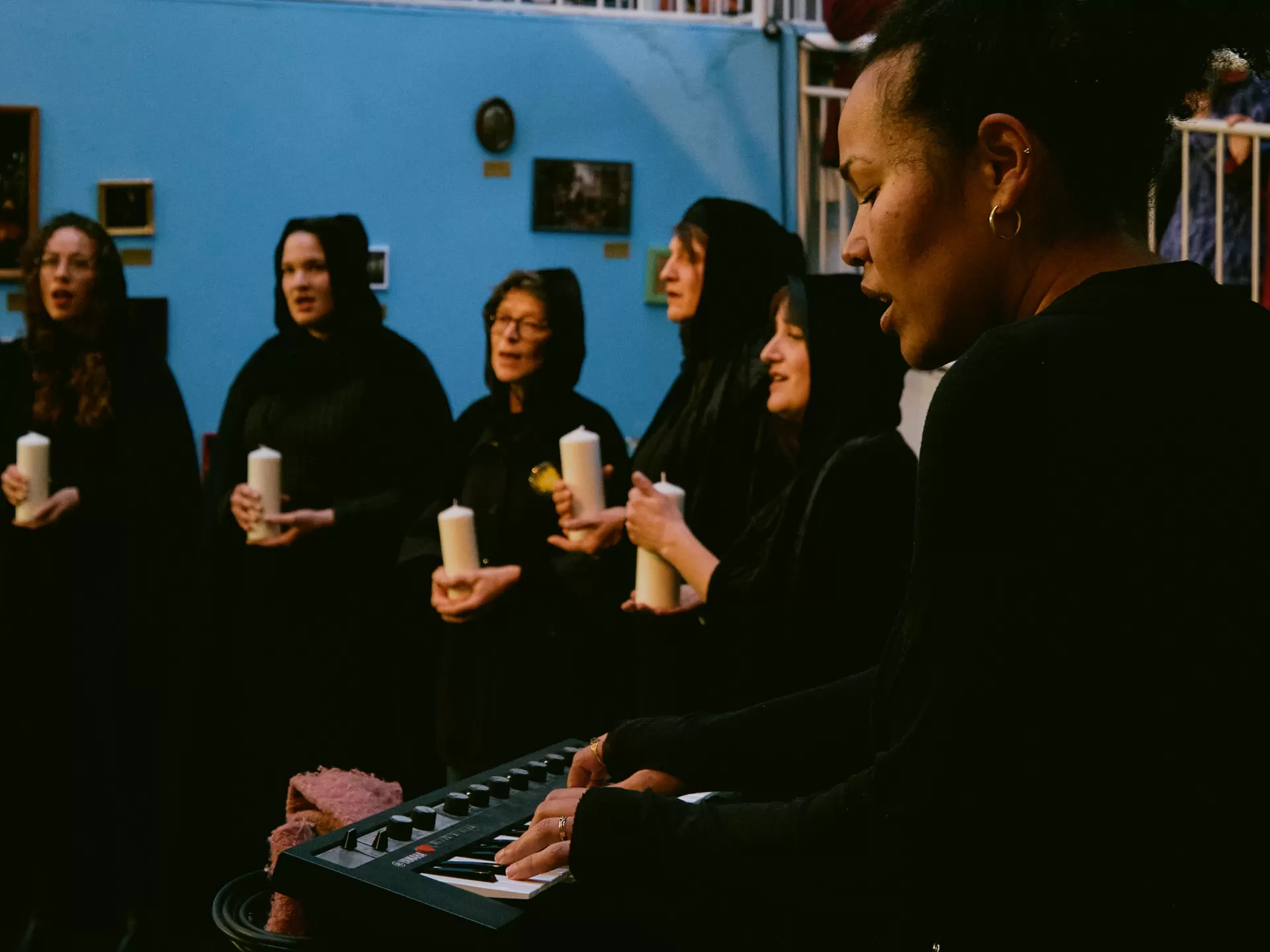 Eine Frau spielt und singt an einem Keyboard. Dahinter stehen die anderen Personen in schwarzen Kutten und mit Kerzen in der Hand.