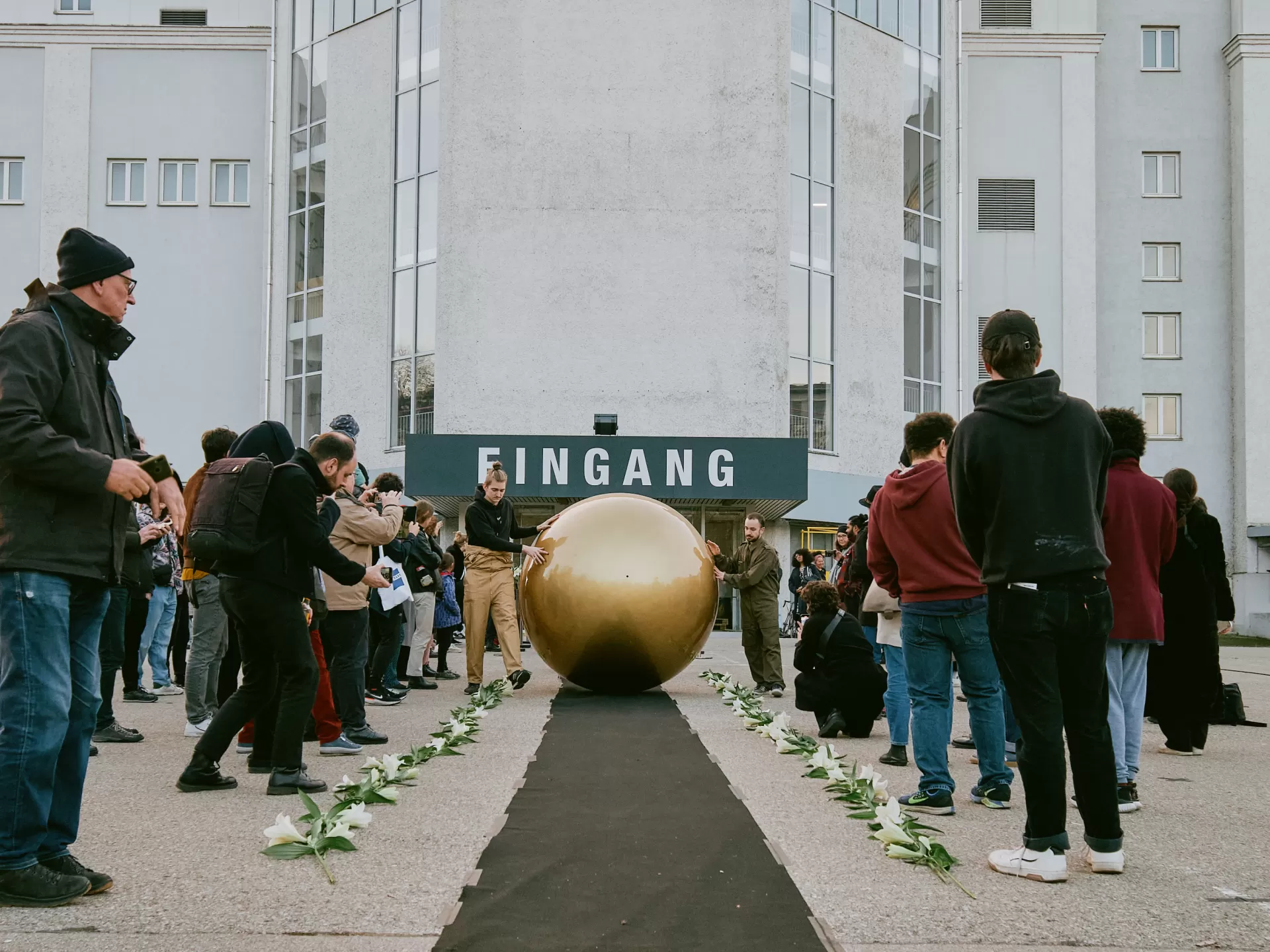 Zwei Männer rollen die goldene Kugel einen schwarzen langen Teppich herab. An den Seiten steht das Publikum und schaut zu.