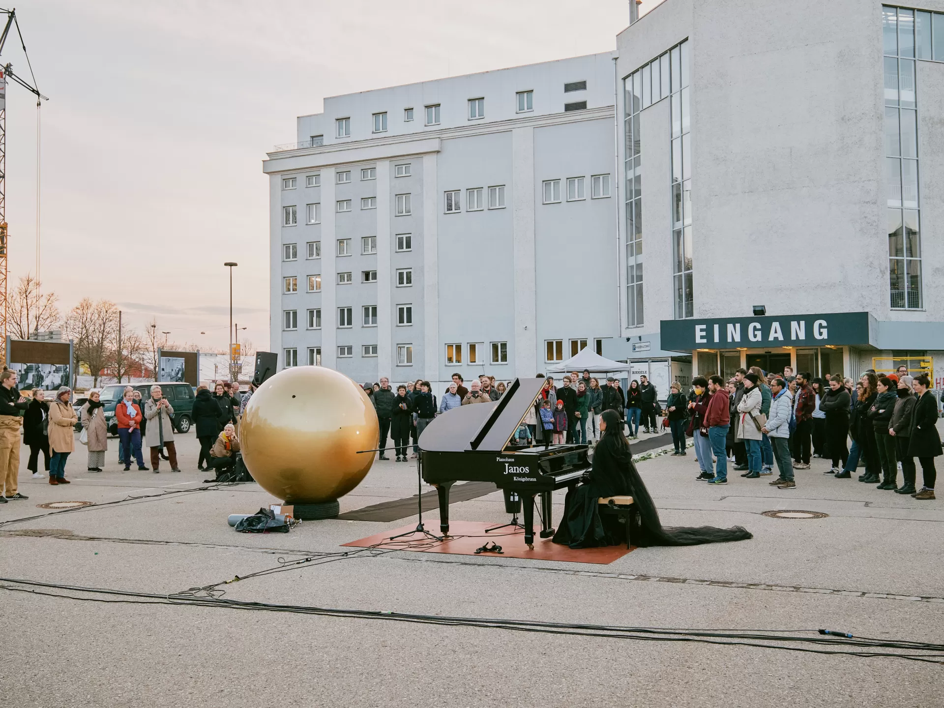 Auf einem Parkplatz hat sich eine Menschenmenge im Halbkreis um das Geschehen gestellt. Die goldene Kugel steht am Ende des schwarzen Teppichs. Daneben sitzt eine junge Frau mit schwarzem Umgang und Teufelshörnern an einem Flügel.