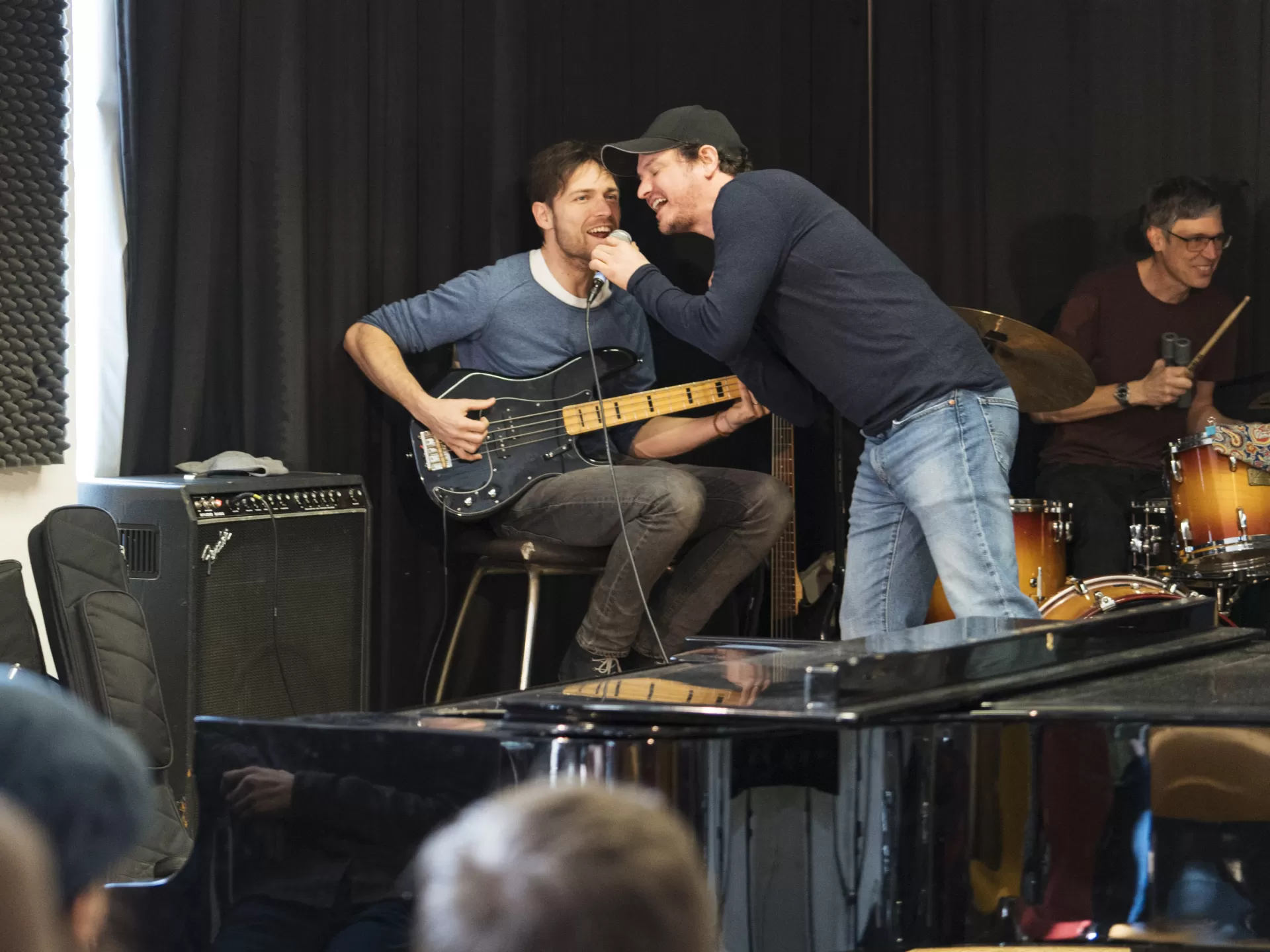 Ein interner Auftritt der Coach-Band. Zwei Coaches singen zusammen in ein Mikrofon. Einer von beiden sitzt auf einem Stuhl und spielt E-Bass.