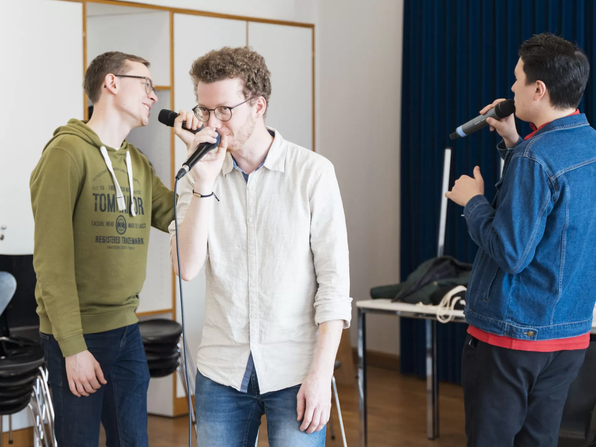 Drei Sänger mit Mikrofonen im Coaching. Die Person in der Mitte schaut nach vorne, die beiden äußeren singen sich gegenseitig an.