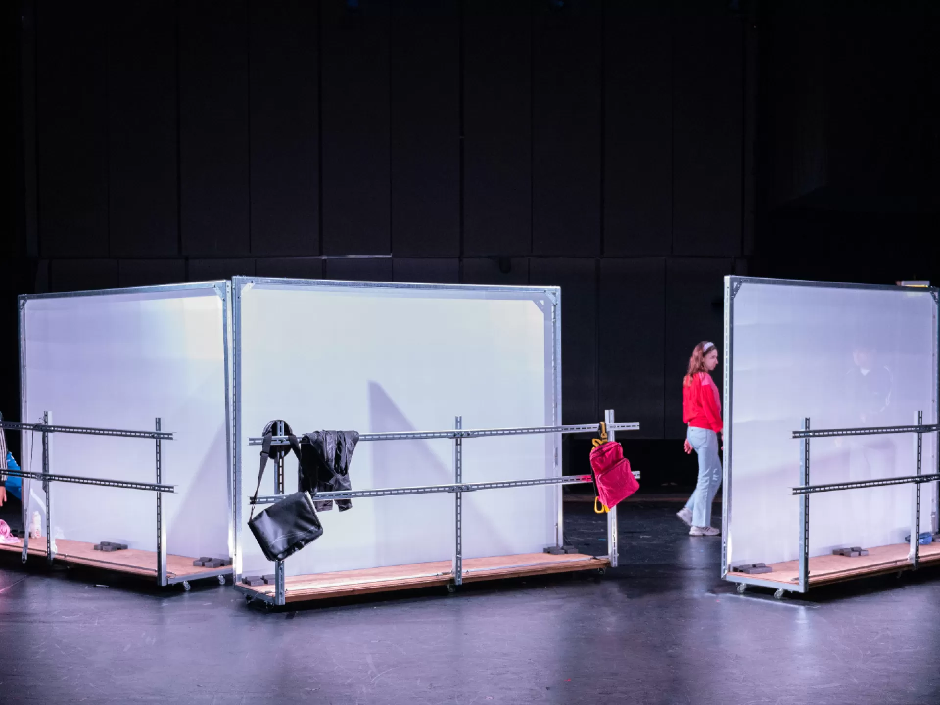 Drei einzelne Stellwände, weiß und leicht transparent, mit jeweils einer Metallbrüstung davor sind schräg gegeneinaner angeordnet, am linken Rand ist ein Mann zu sehen, in der Mitte eine Frau. An den Metallbrütungen hängen Kleidungsstücke und Taschen.