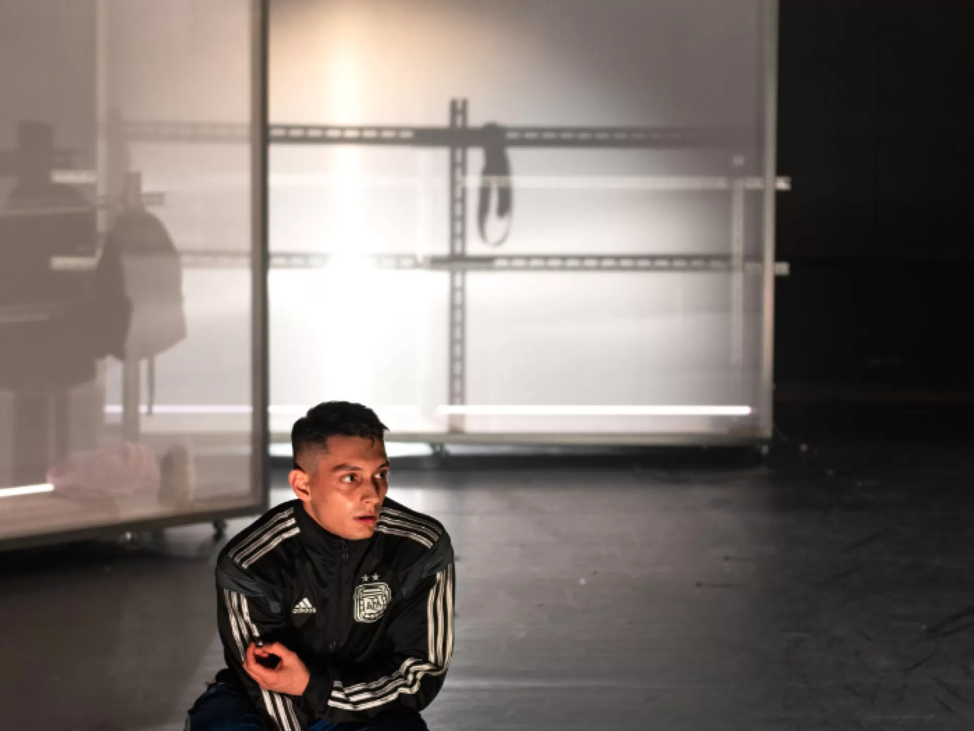 Ein Ausschnitt eines Bühnenraumes, im Hintergrund zwei weiße, halbtransparente Stellwände mit Metallbrüstung davor. Im Vordergrund hockt ein Mann in Turnschuhen und Adidas Trainingsanzug, er schaut fragend nach rechts.